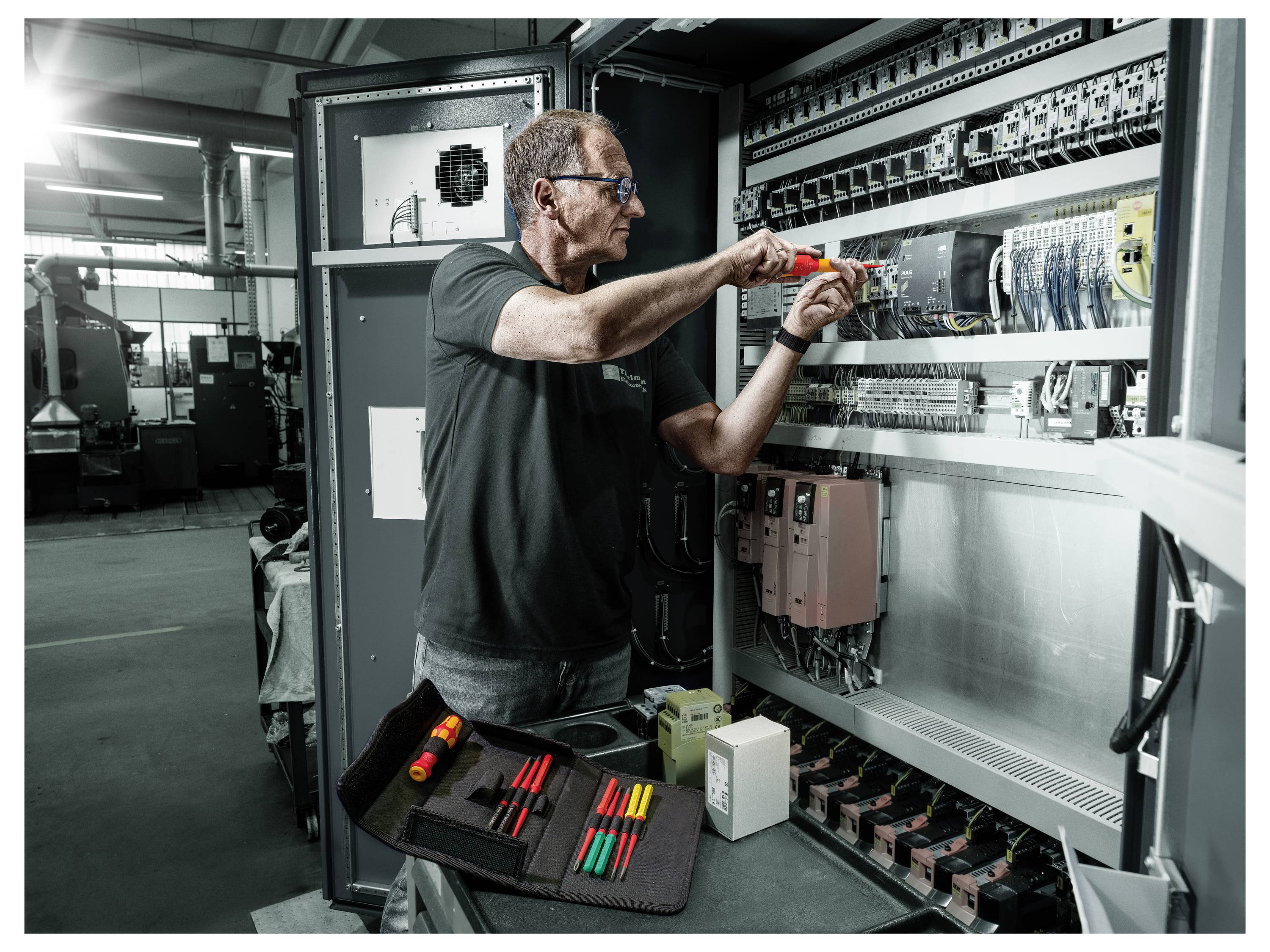 Techniker, der mit einem Schraubendreher an einem offenen elektrischen Schaltschrank in einer Industrieanlage arbeitet, mit in der Nähe angeordneten Werkzeugen.