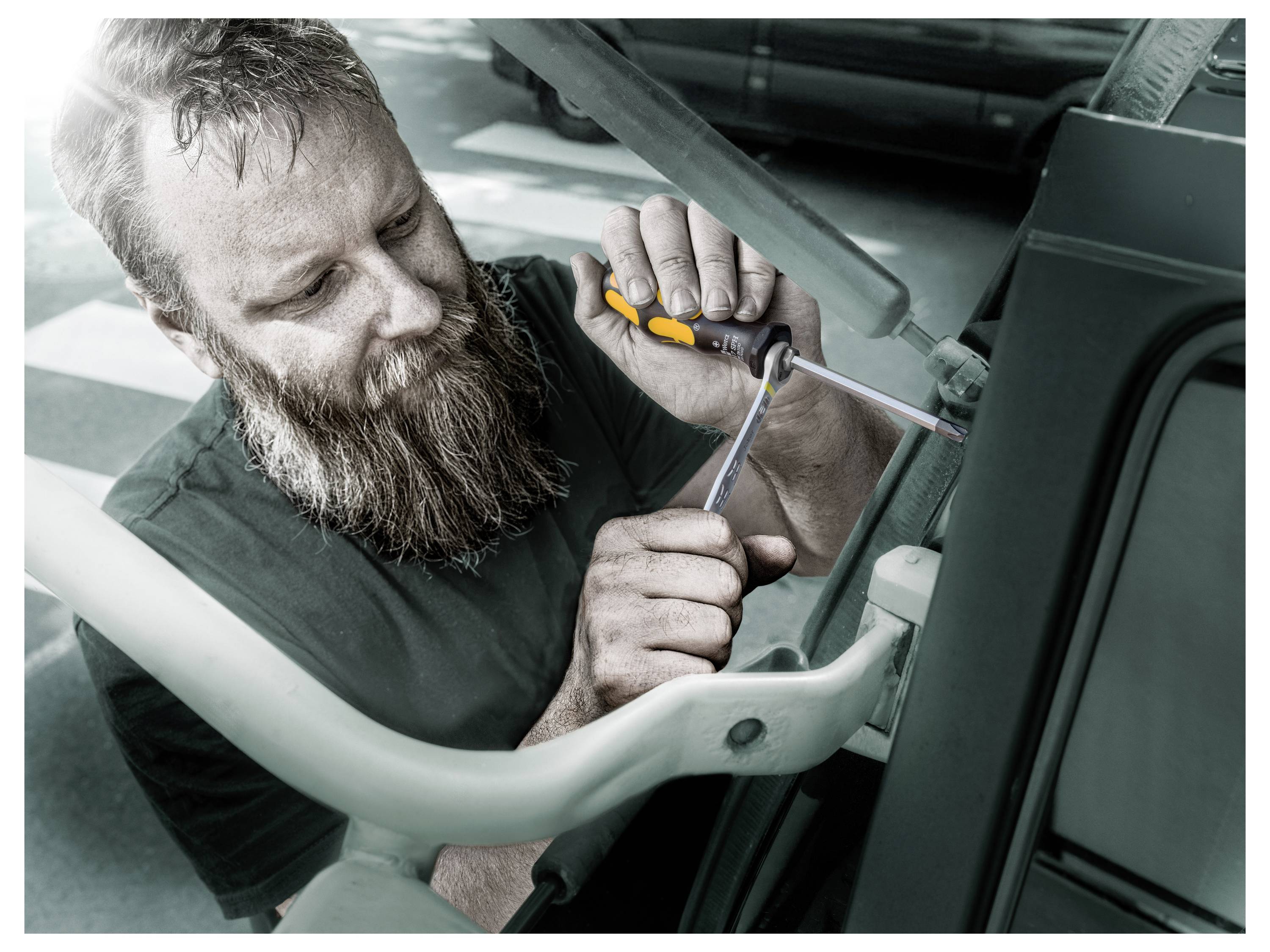 Mechaniker, der mit einem Schraubendreher ein Autoteil unter der Motorhaube festzieht, neben dem Fahrzeug in einer Werkstatt arbeitet.