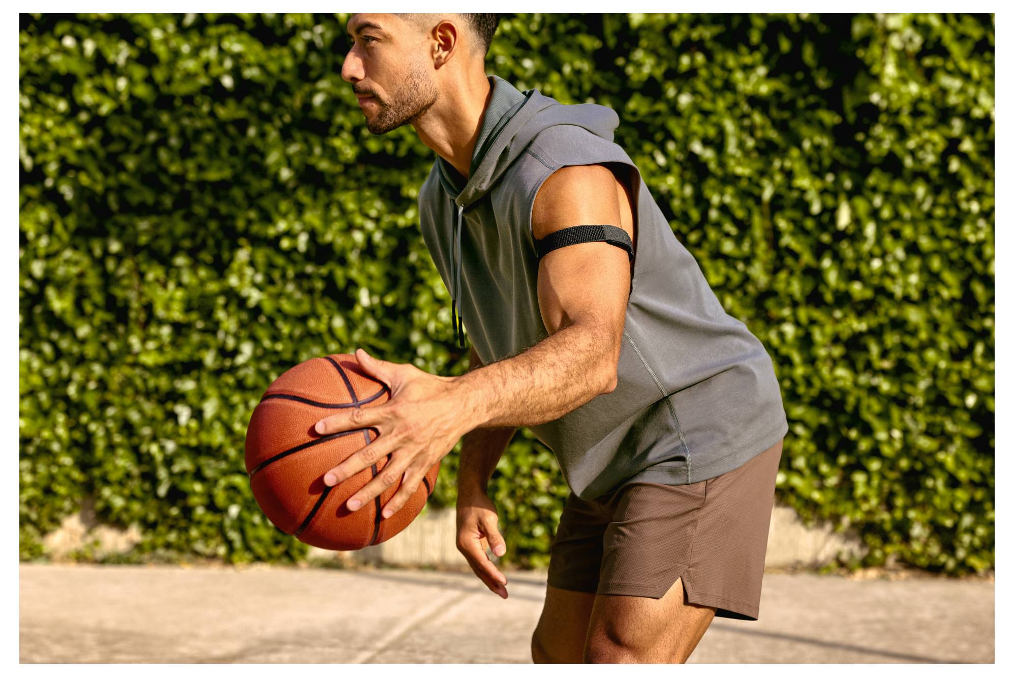 Ein Mann dribbelt einen Basketball im Freien auf einem sonnigen Platz, trägt ein ärmelloses Hoodie und Shorts und steht in einer Bereitschaftsposition.