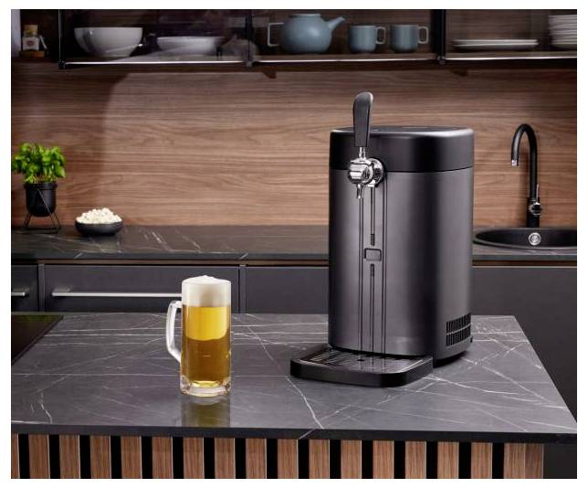 Bierabfüller auf einer Küchenarbeitsplatte neben einem gefüllten Bierkrug, mit einem Waschbecken und Regalen im Hintergrund.