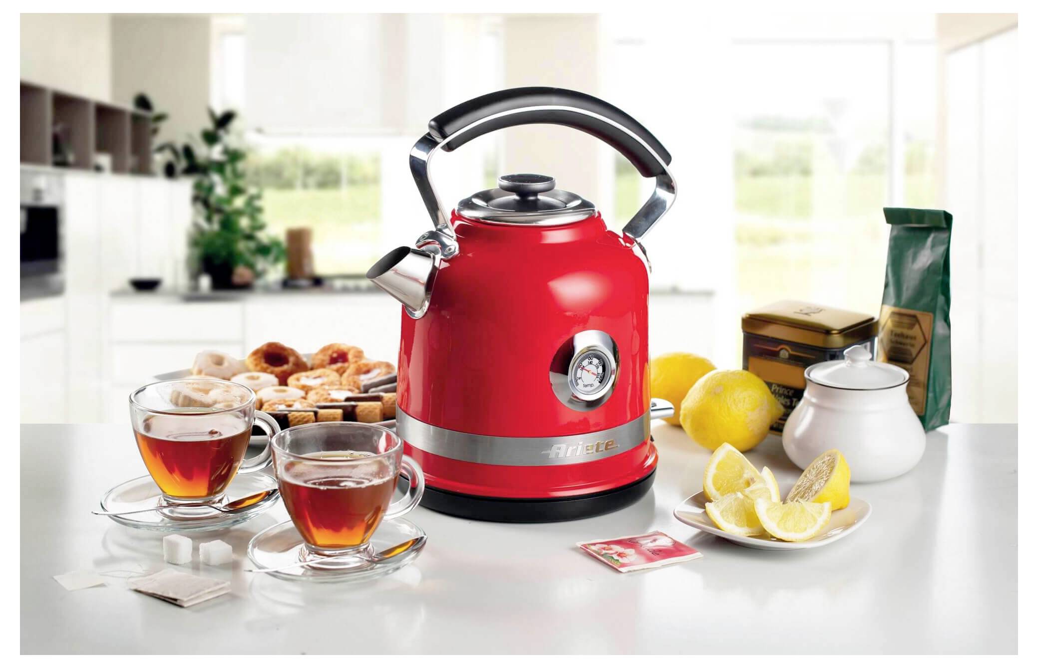 Roter Wasserkocher auf einer Küchentheke mit Teetassen, Donuts, Zitronen und einer Zuckerdose, was auf eine Tee- oder Frühstückseinrichtung hindeutet.