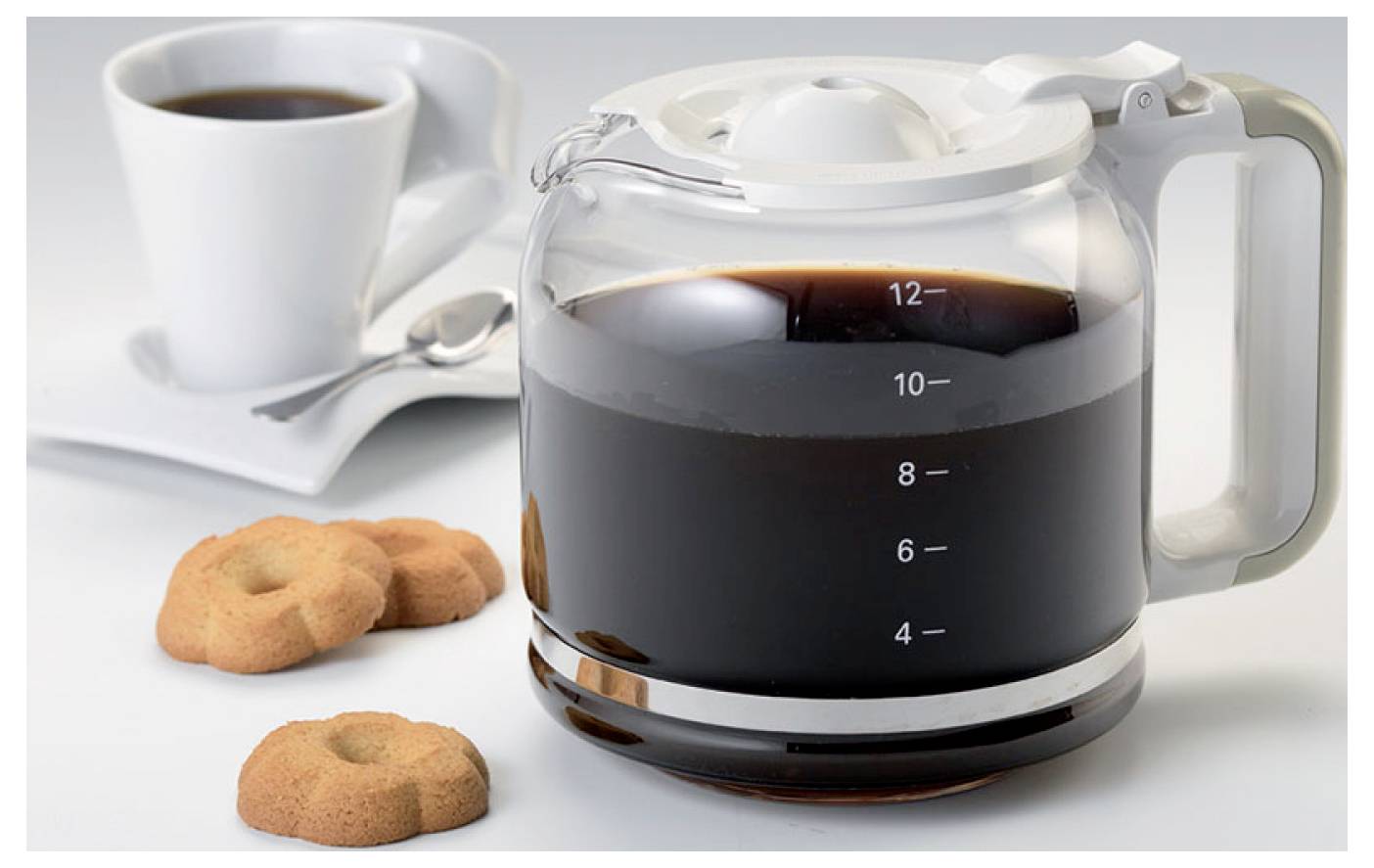 Glaskaffeekanne gefüllt mit gebrühtem Kaffee neben einer Tasse und Untertasse, mit Keksen im Vordergrund.