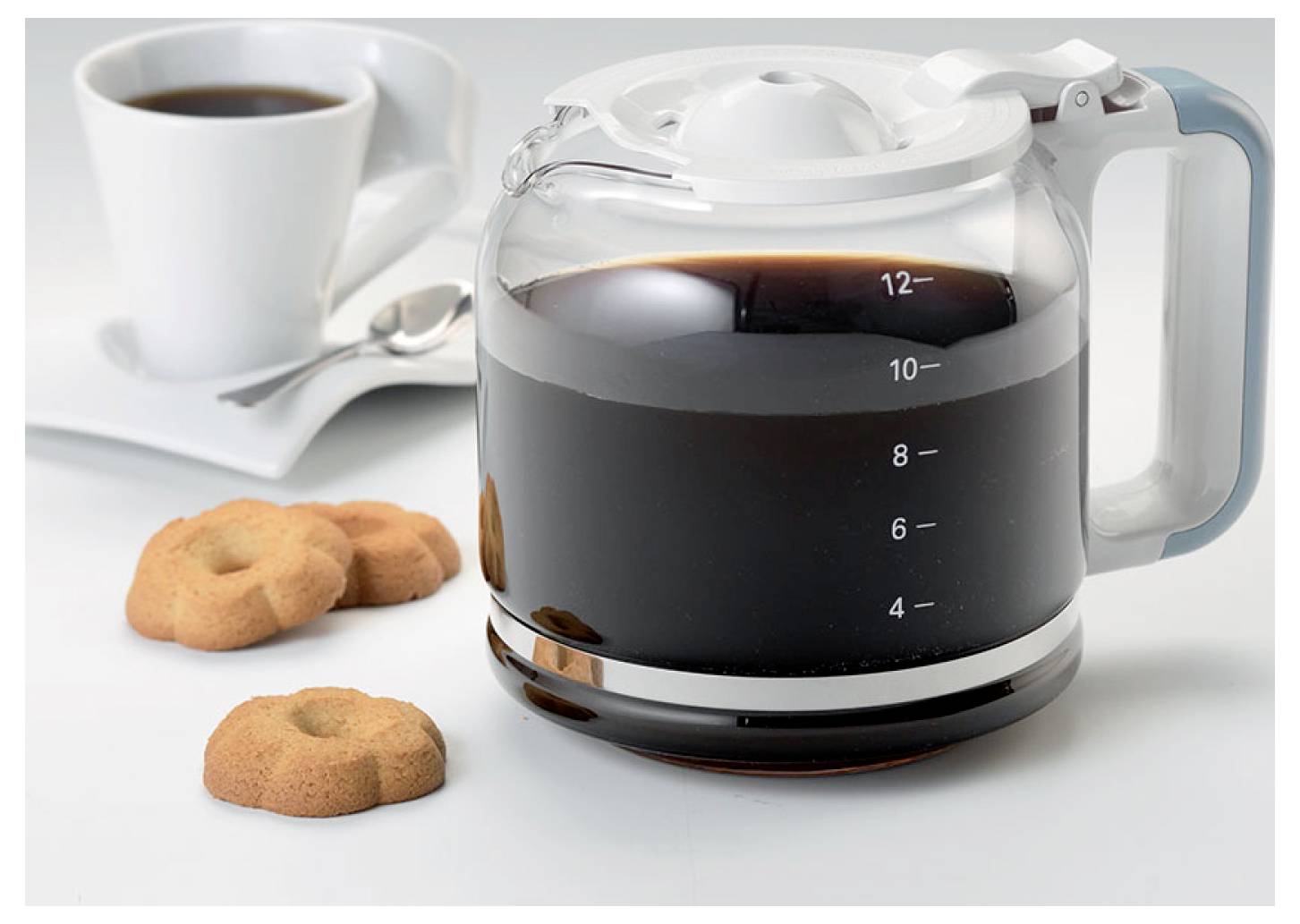 Eine Glaskaffeekanne gefüllt mit Kaffee steht neben einer weißen Tasse, einem Löffel und Keksen auf einer hellen Oberfläche.