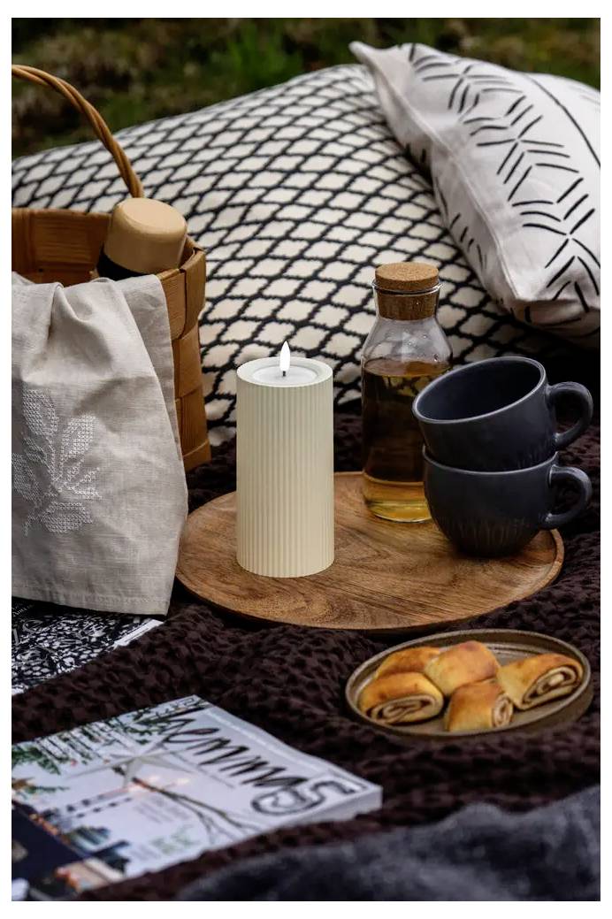Picknick-Aufbau mit einem Korb, einer Kerze, Kaffeetassen, einem Glas Sirup, Backwaren, einer Zeitschrift und Kissen auf einer gemusterten Decke im Freien.