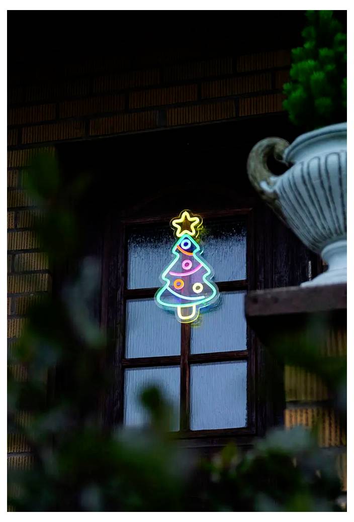 Ein leuchtendes Neon-Weihnachtsbaumsschild strahlt in einem Fenster, mit einem Stern oben drauf und einem großen Blumentopf, der auf der rechten Seite teilweise sichtbar ist.