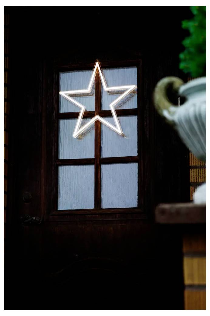 Ein leuchtender, sternförmiges Licht, das in einem Fenster einer dunkelbraunen Holztür hängt, mit einer unscharfen Pflanze oder einer Vase im Vordergrund.