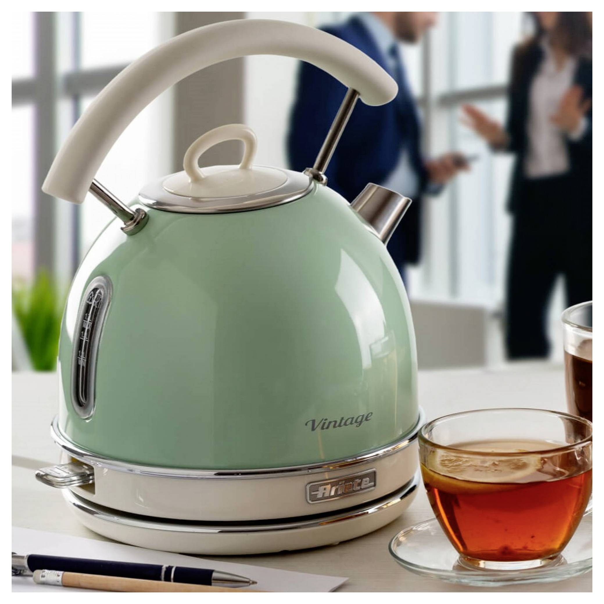 Minzgrüner vintage Wasserkocher auf einem Tisch neben einer Tasse Tee und einem Stift, mit unscharfen Menschen im Hintergrund.