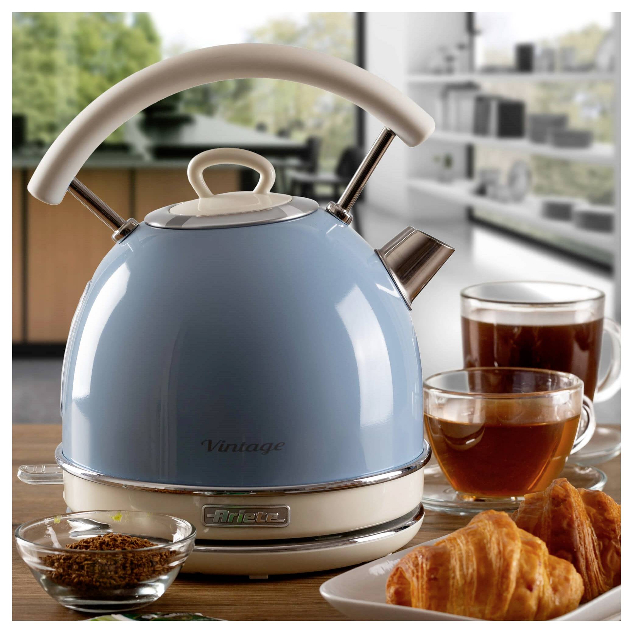 Blauer Wasserkocher auf einem Tisch mit Tee und Croissants in einer hellen Küche.