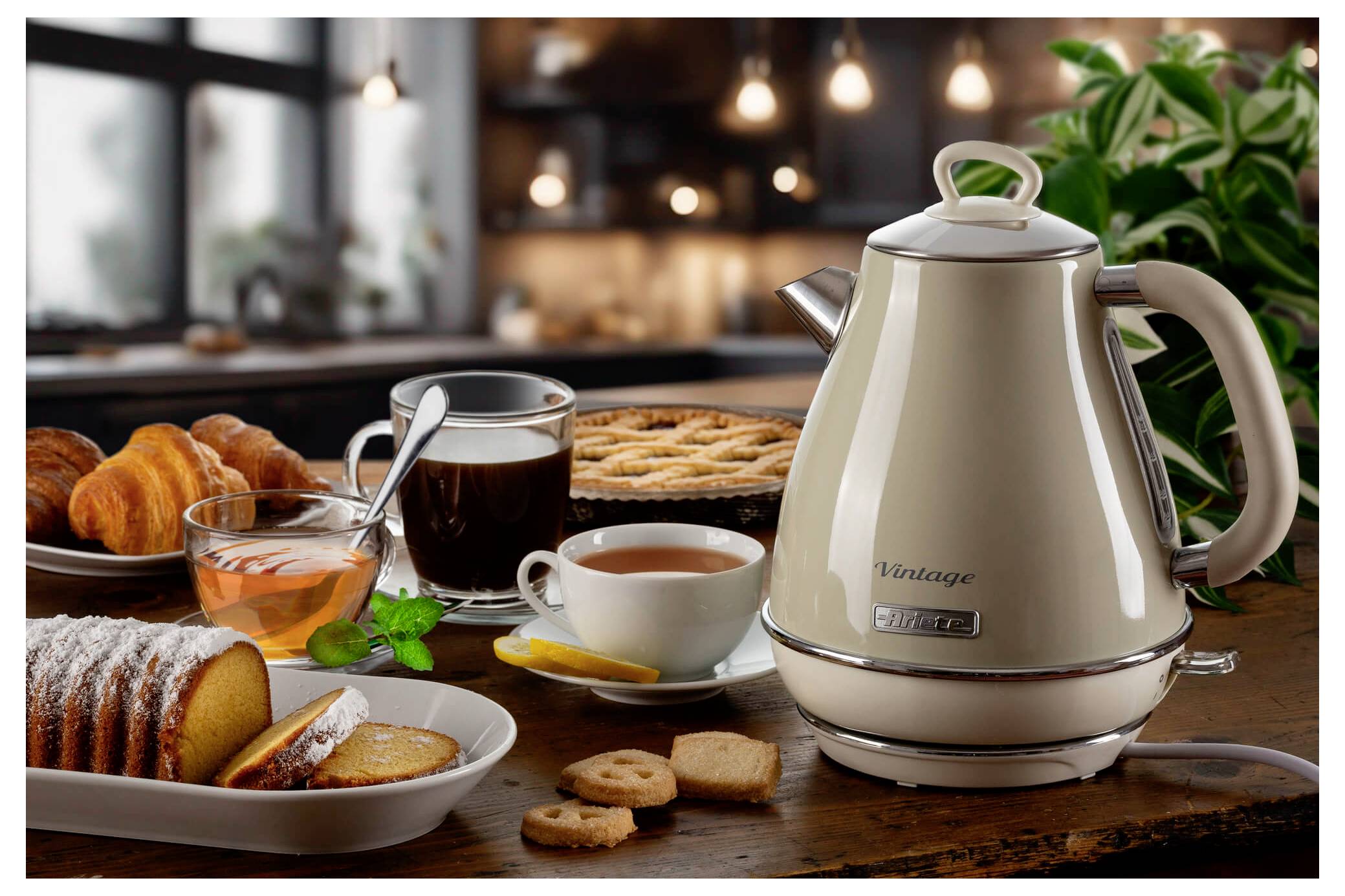 Küchentheke mit elektrischem Vintage-Wasserkocher, Kaffeetassen, Tee, Croissants, Kuchen, Keksen und einer Topfpflanze im Hintergrund.