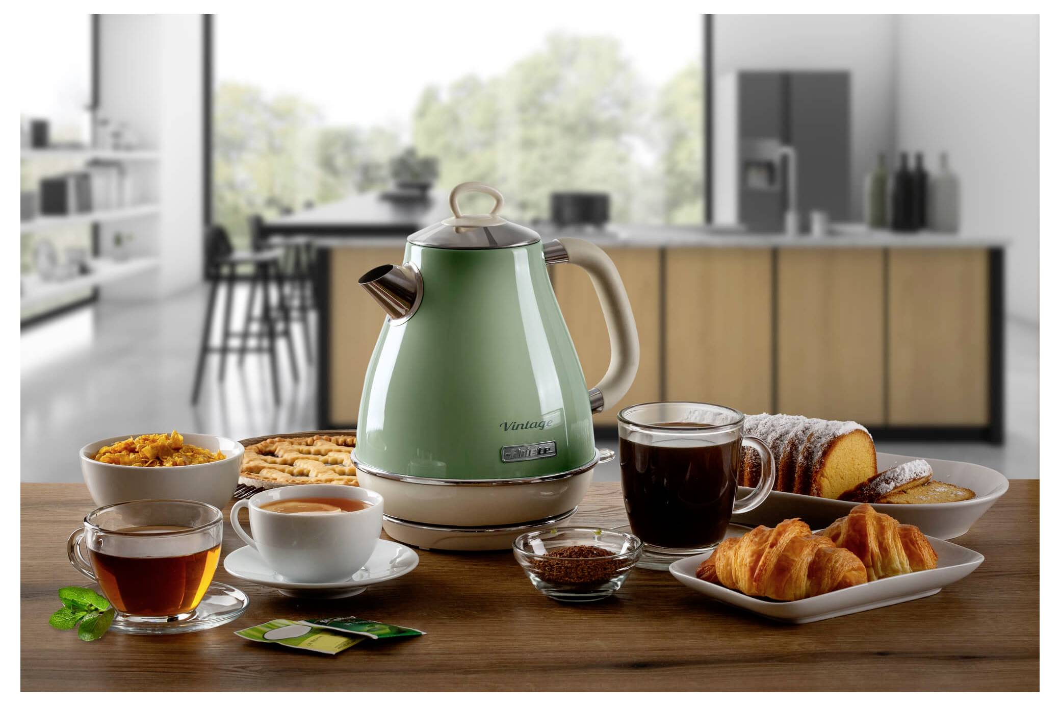 Frühstücksauslage auf einem Holztisch mit einem grünen Elektrokessel, Tee, Kaffee, Gebäck und Marmelade in einer modernen Küche