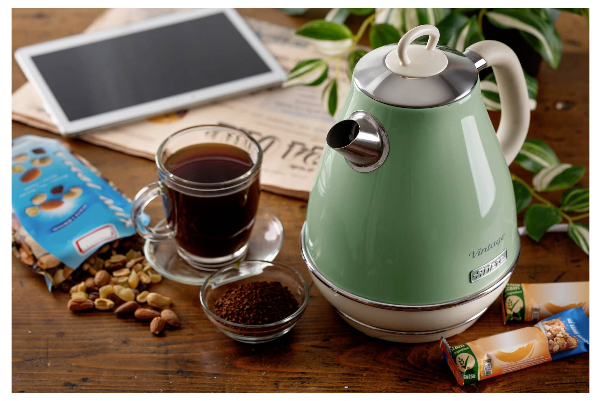 Grüner Wasserkocher neben einem Glas Tee auf einem Holztisch mit Kaffeebohnen, Tablet, Nüssen und Müsliriegeln