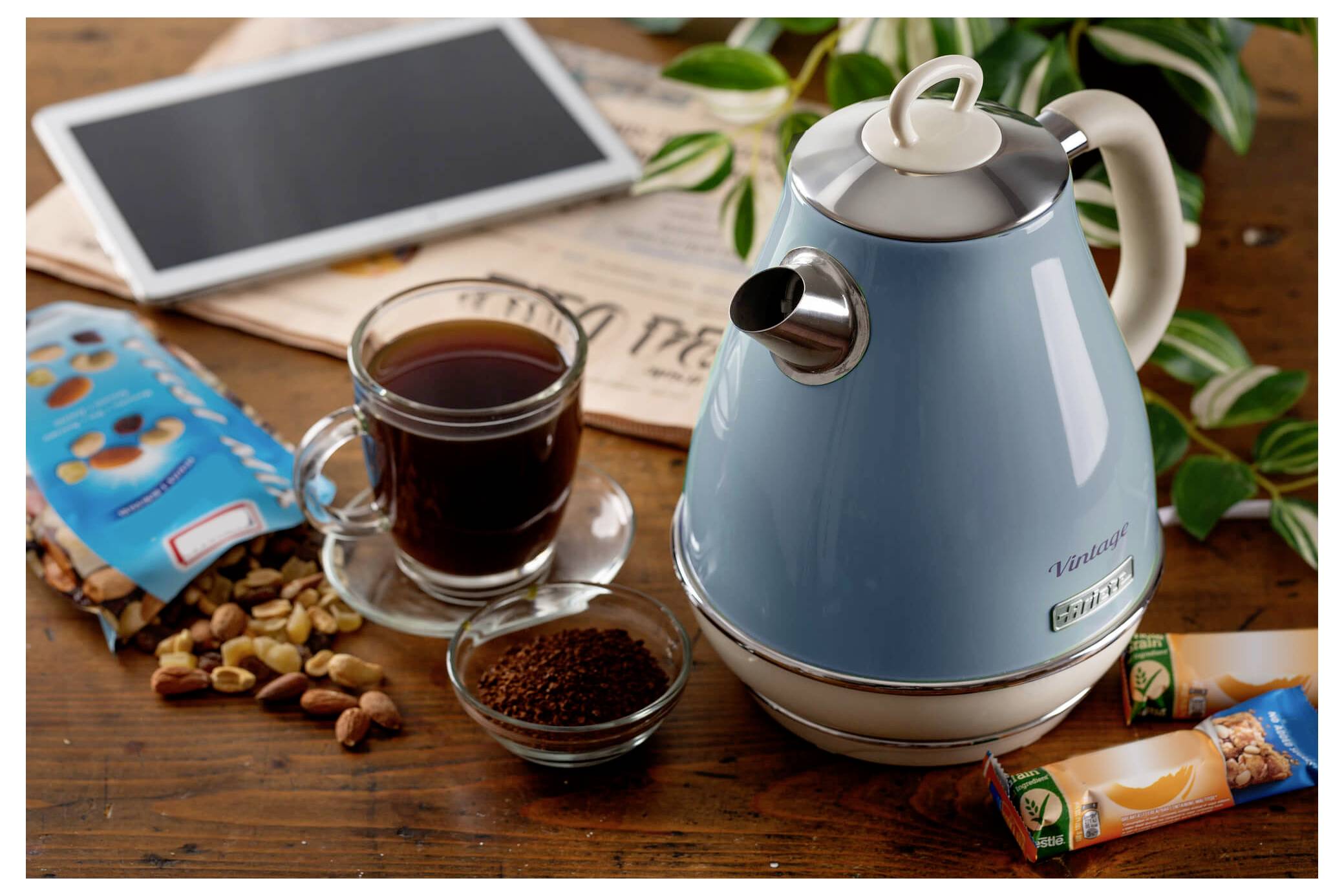 Blauer Wasserkocher auf einem Holztisch neben einer Tasse Kaffee, einer kleinen Schüssel mit Kaffeepulver, Snack-Verpackungen, einem Tablet und Pflanzenblättern.