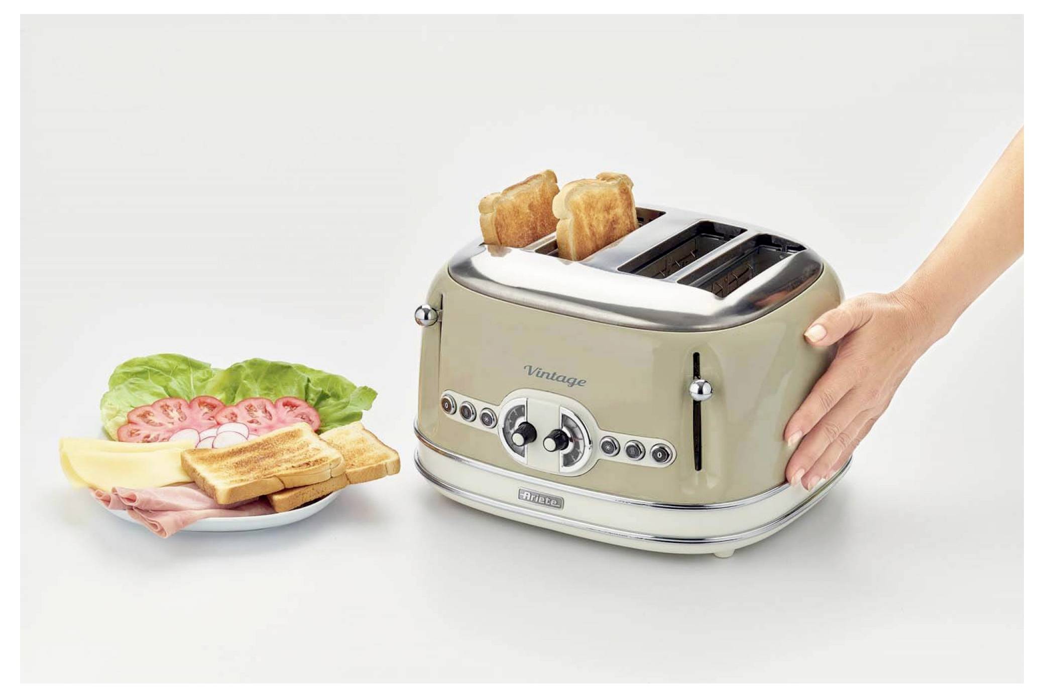 Eine Hand legt Toast in einen Vintage-Toaster für vier Scheiben neben einen Teller mit Brot, Aufschnitt, Salat und Tomate auf weißem Hintergrund.