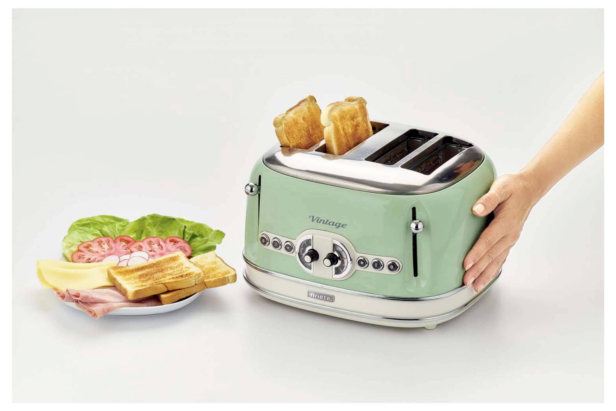 Hand, die Toast in einen minzgrün gefärbten Vintage-Vier-Schlitz-Toaster neben einen Teller mit getoasteten Toastbrot mit Schinken, Salat und Tomate legt.