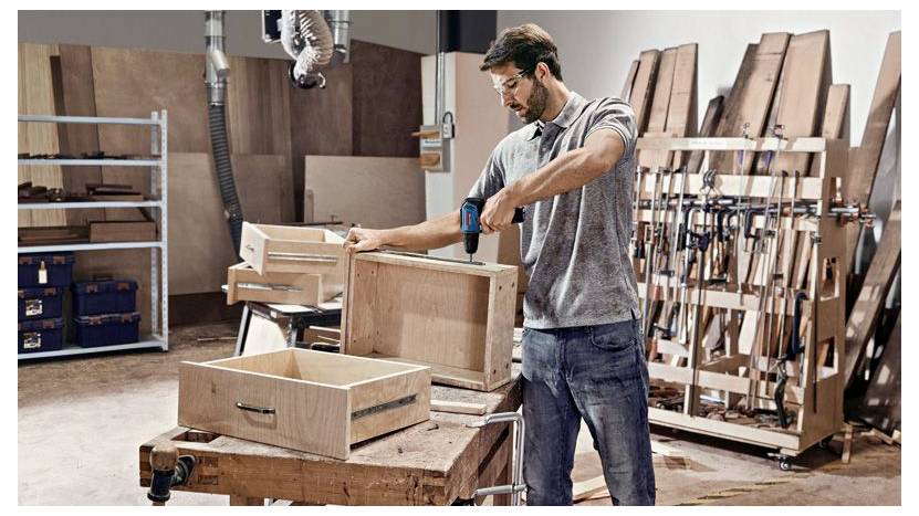 Ein Mann montiert eine hölzerne Schublade mit einem Bohrer in einer Tischlerwerkstatt, umgeben von Holz, Werkzeugen und Werkbänken.