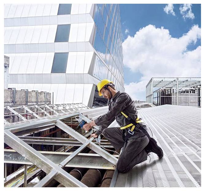 Bauarbeiter in gelbem Schutzhelm, der ein Metallgerüst auf einem Dach neben einem modernen Glasgebäude sichert.
