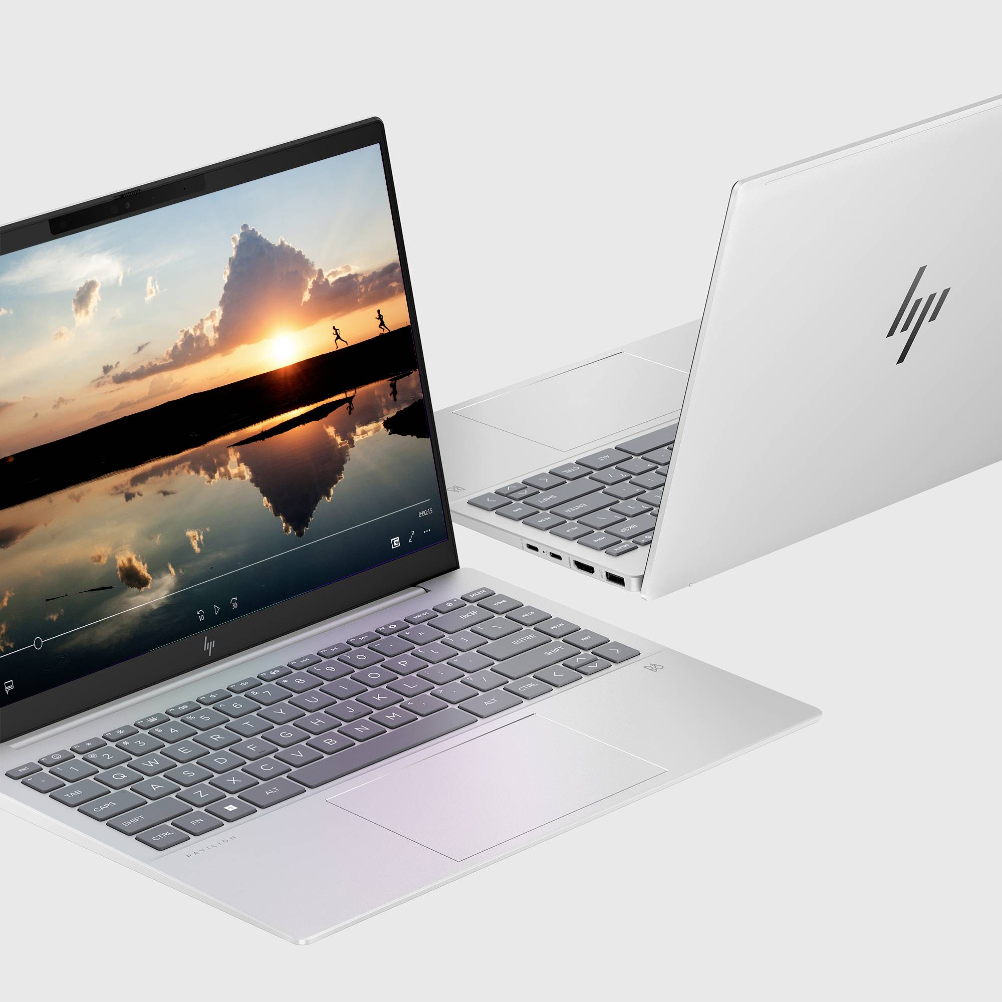 Zwei silberne HP-Laptops in geöffnetem Zustand in verschiedenen Winkeln dargestellt, einer zeigt ein Sonnenuntergang-Video auf dem Bildschirm und der andere zeigt das HP-Logo auf dem Deckel.