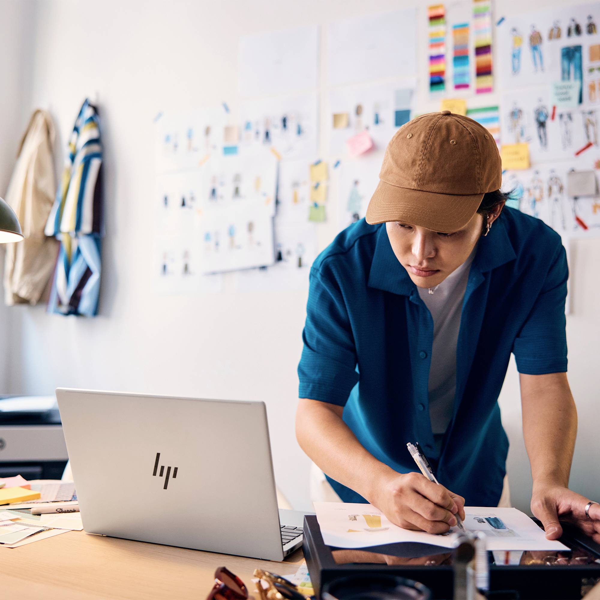 Eine Person in einer braunen Kappe und blauem Hemd skizziert auf Papieren an einem Schreibtisch neben einem offenen Laptop in einem Designstudio.