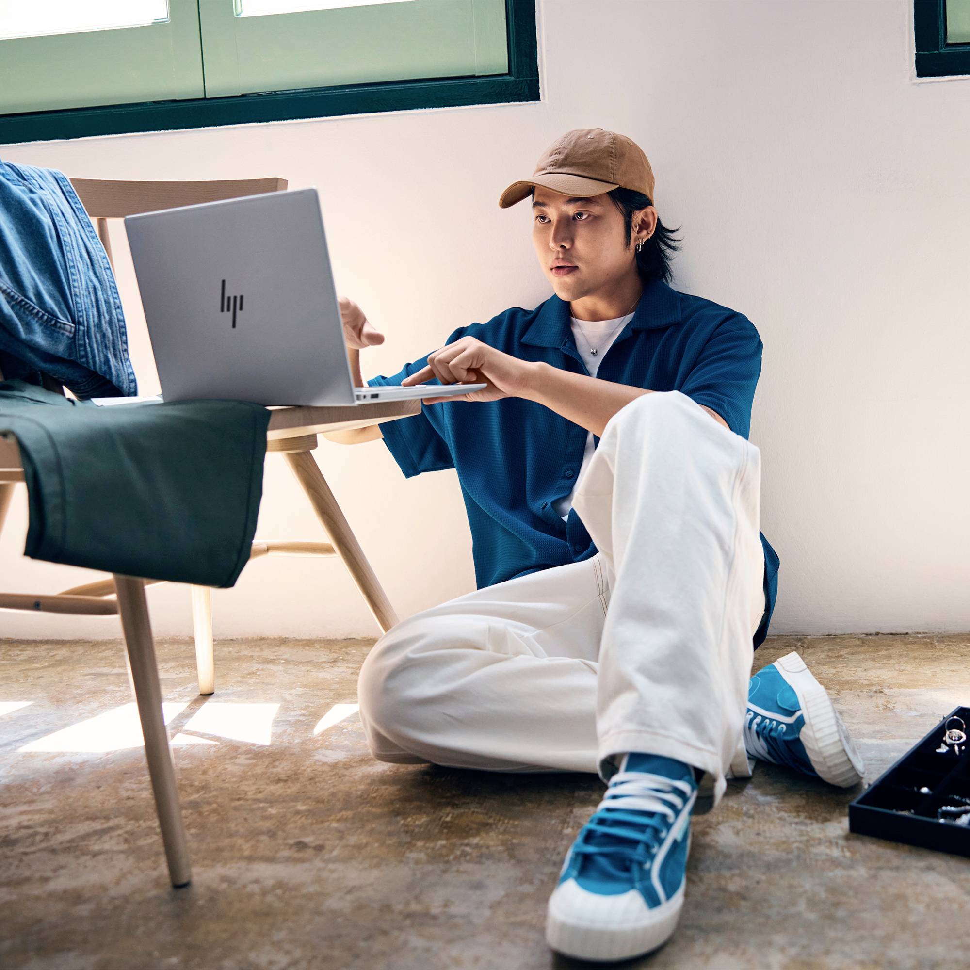 Junger Mann, der neben einem Tisch auf dem Boden sitzt, einen Laptop benutzt und sich nach einem Objekt ausstreckt, in einem hellen Raum.