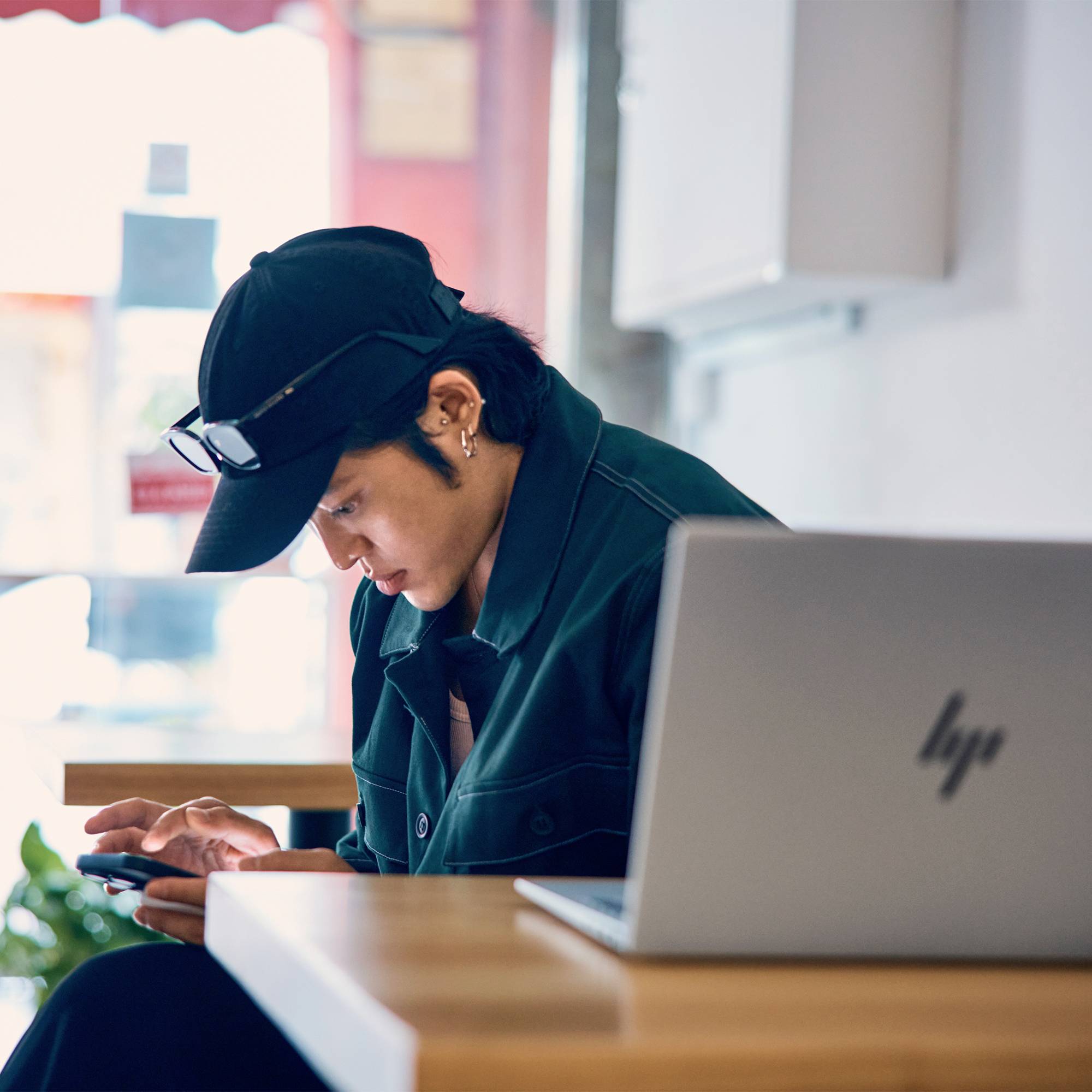 Person in einer Mütze und Brille, die ein Smartphone an einem Tisch neben einem aufgeklappten HP-Laptop in einem hellen Café oder Arbeitsbereich nutzt.