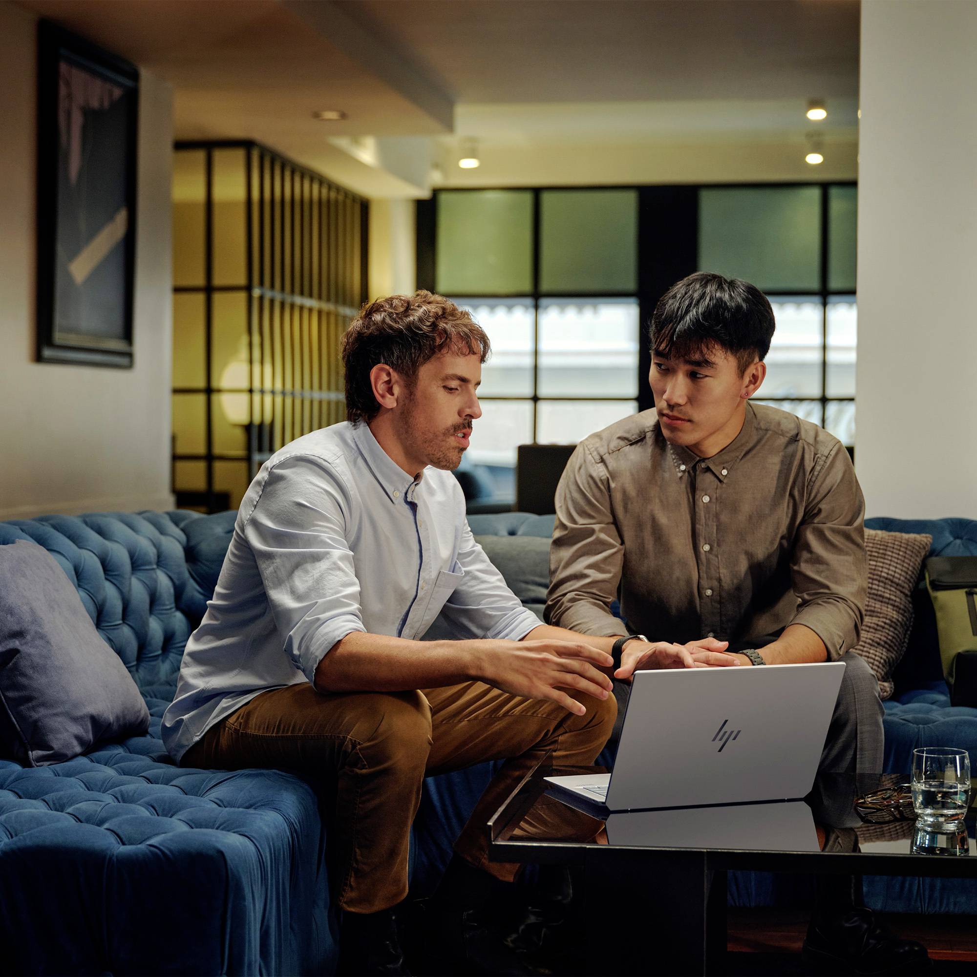 Zwei Männer diskutieren etwas auf einem Laptop, während sie auf einem blauen Sofa in einem modernen Wohnzimmer sitzen.