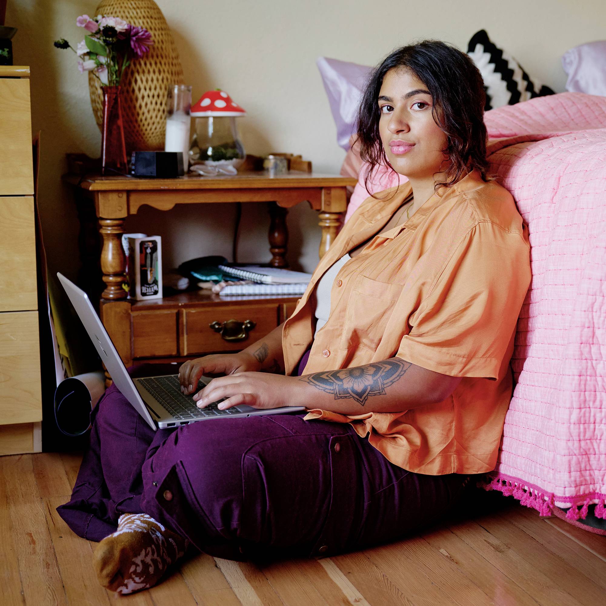 Frau, die auf dem Boden neben einem Bett sitzt und einen Laptop benutzt, in einem gemütlichen Schlafzimmer mit einem Holzschreibtisch und einer Lampe hinter ihr.