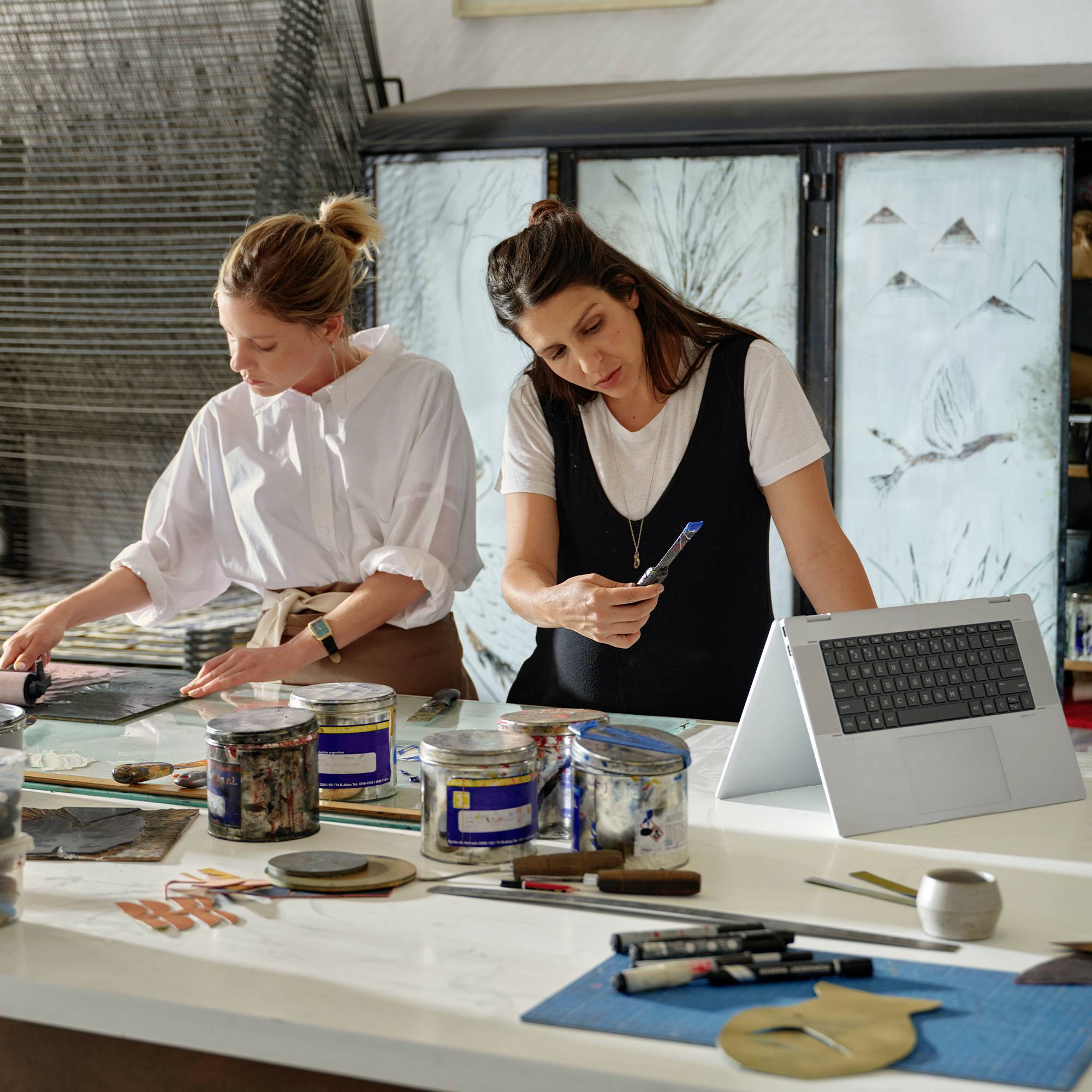 Zwei Frauen arbeiten an einem Tisch in einem Kunstatelier und malen sowie überprüfen Materialien, wobei Farbdosen, Pinsel, ein Laptop und Kunstbedarf sichtbar sind.