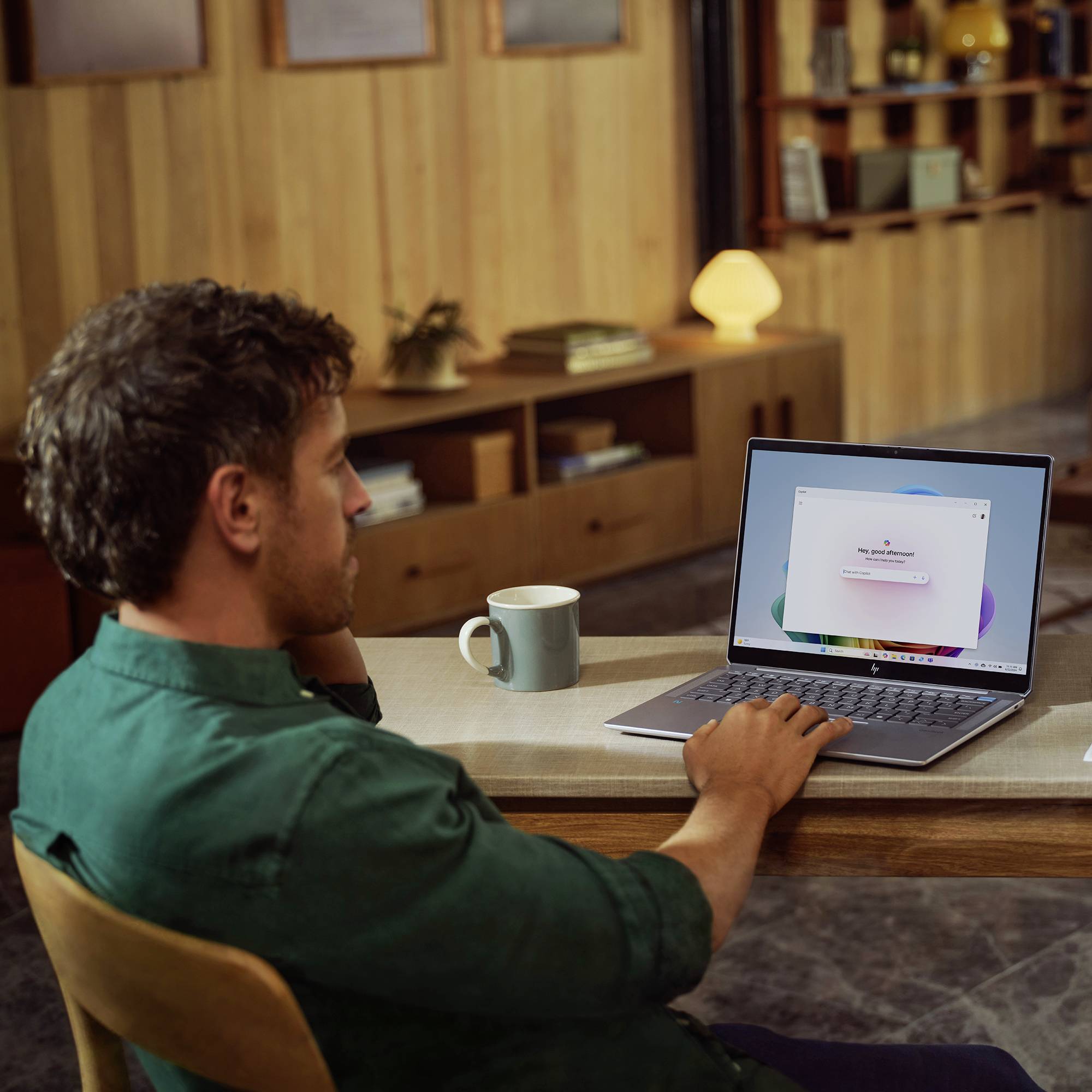Ein Mann sitzt mit einem Laptop an einem Tisch in einem gemütlichen Zimmer; neben ihm steht eine Tasse und der Bildschirm zeigt ein Softwarefenster.