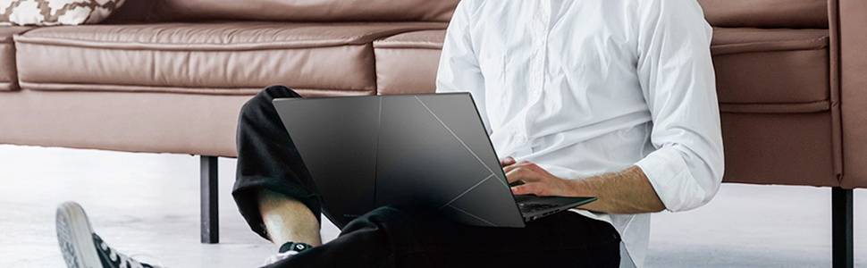 Eine Person sitzt auf dem Boden vor einem braunen Sofa und arbeitet oder tippt auf einem Laptop im Wohnzimmer.
