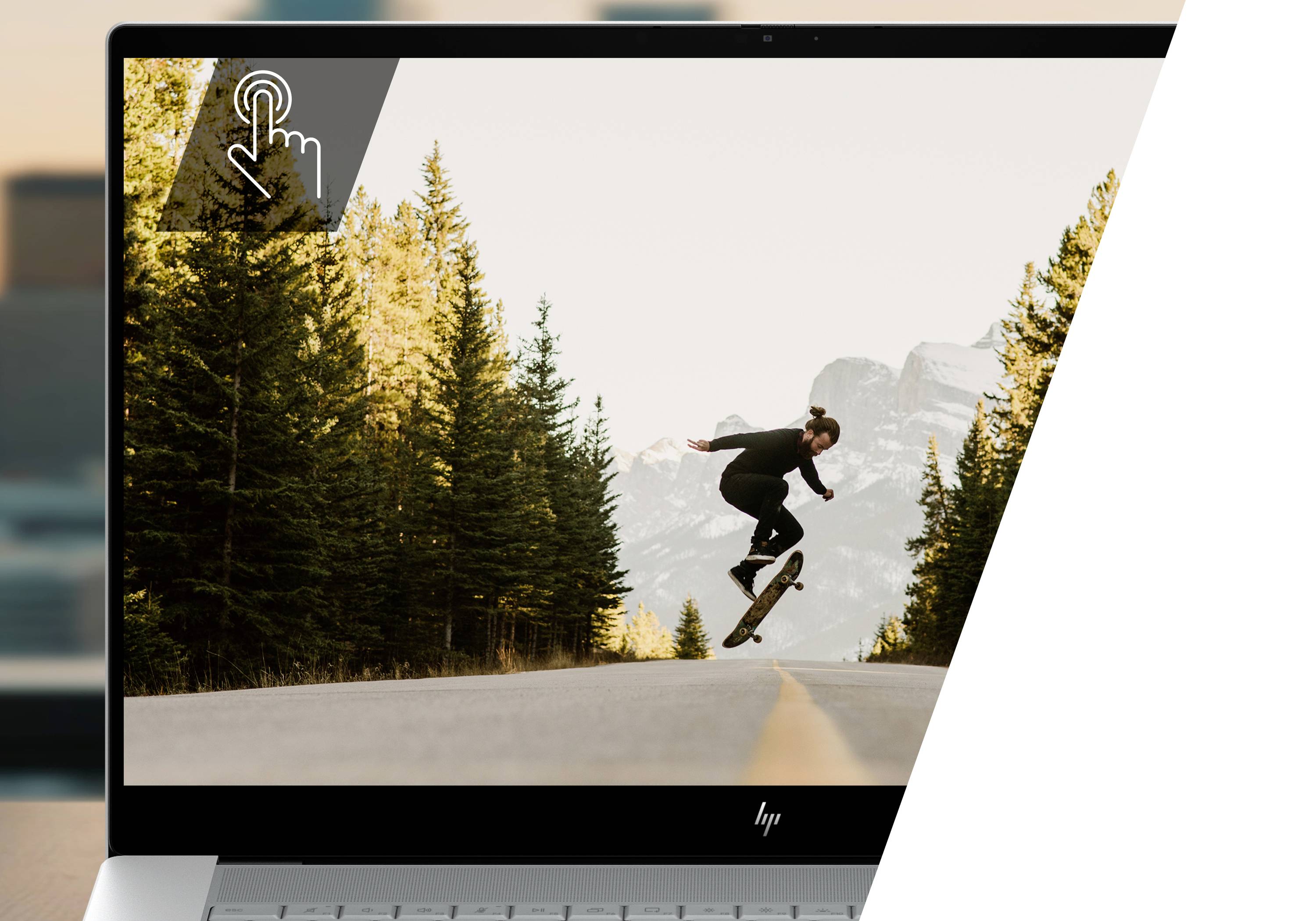 Laptop-Bildschirm zeigt einen Skateboardfahrer bei einem Sprung auf einer Bergstraße neben Kiefern, mit einem Touch-Gesten-Symbol oben links.