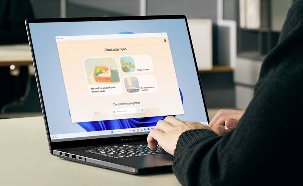 Eine Person tippt auf einem Laptop und zeigt einen Windows-ähnlichen Startbildschirm mit „Guten Nachmittag" und vorgeschlagenen Karten.