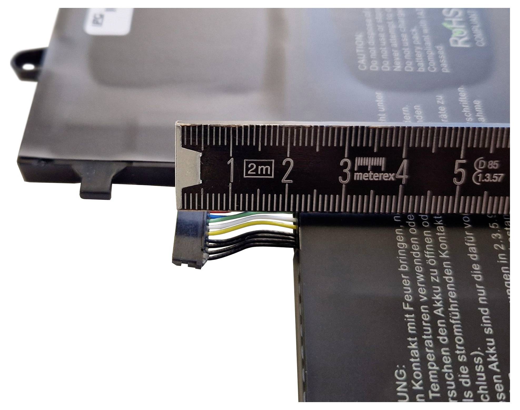 Ein Lineal misst die Dicke eines schwarzen Batteriepakets mit freigelegten mehrfarbigen Drähten und gedrucktem Warntext.