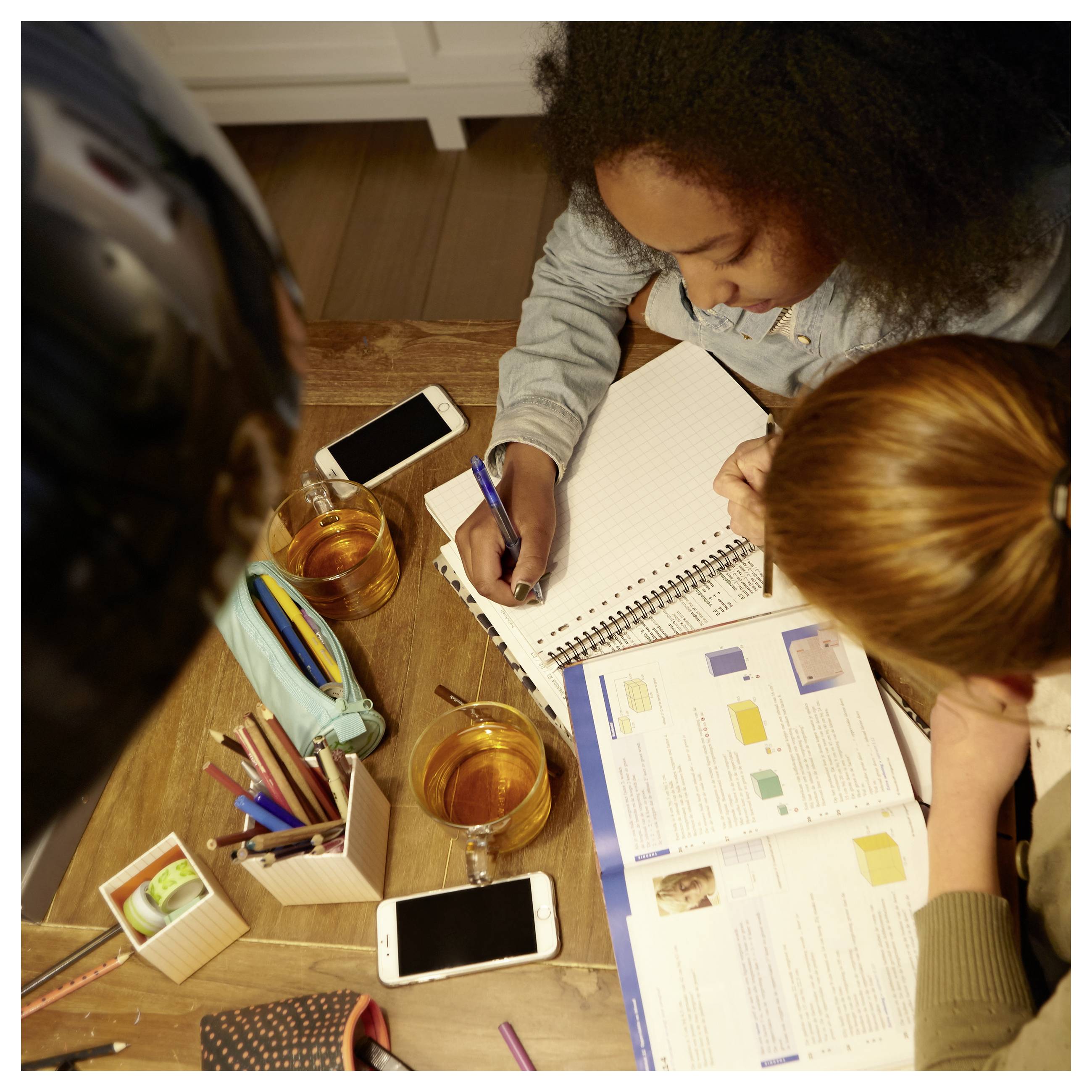 Zwei Kinder arbeiten zusammen an einem Tisch, schreiben in einem Notizbuch und studieren ein Arbeitsbuch mit Getränken, Stiften und Telefonen in der Nähe.