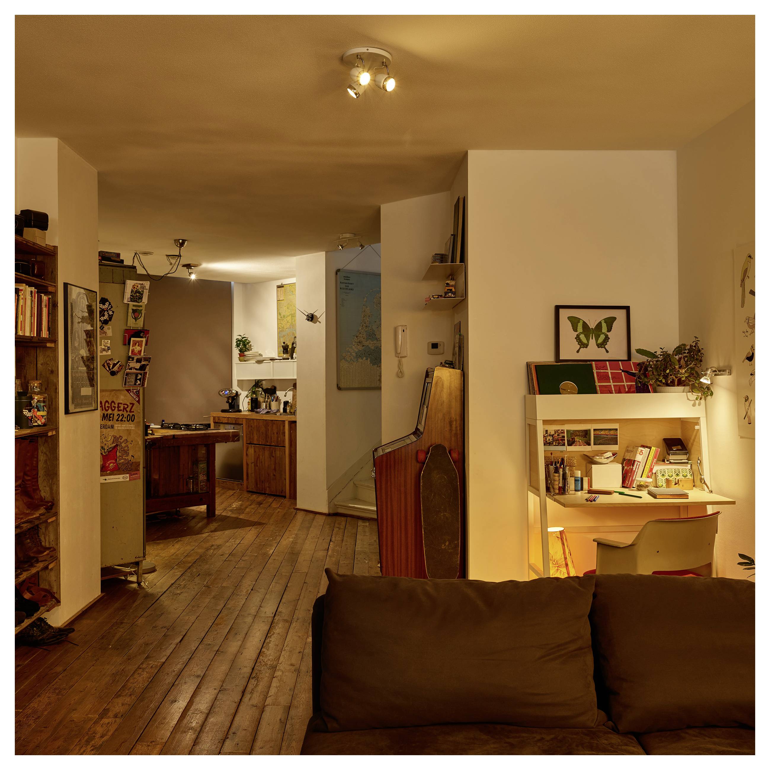 Warmly beleuchtetes Wohnzimmer mit Holzböden, Sofa, Schreibtischecke, Bücherregalen und Küchenbereich im Hintergrund.