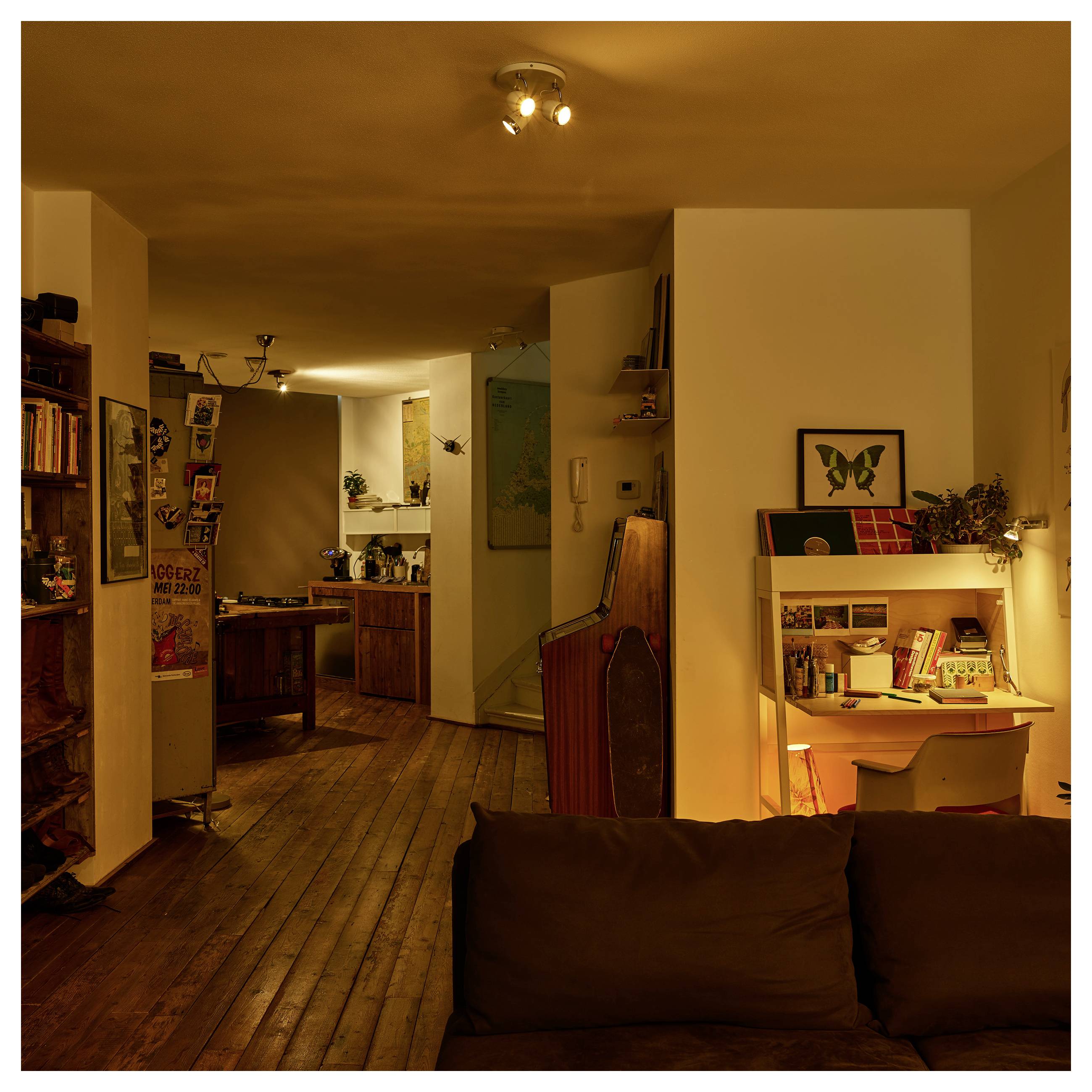 Warm beleuchtetes Apartmentinterieur mit Holzböden, einem dunklen Sofa im Vordergrund, einer kleinen Schreibtischnische auf der rechten Seite und einer Küche im Hintergrund.