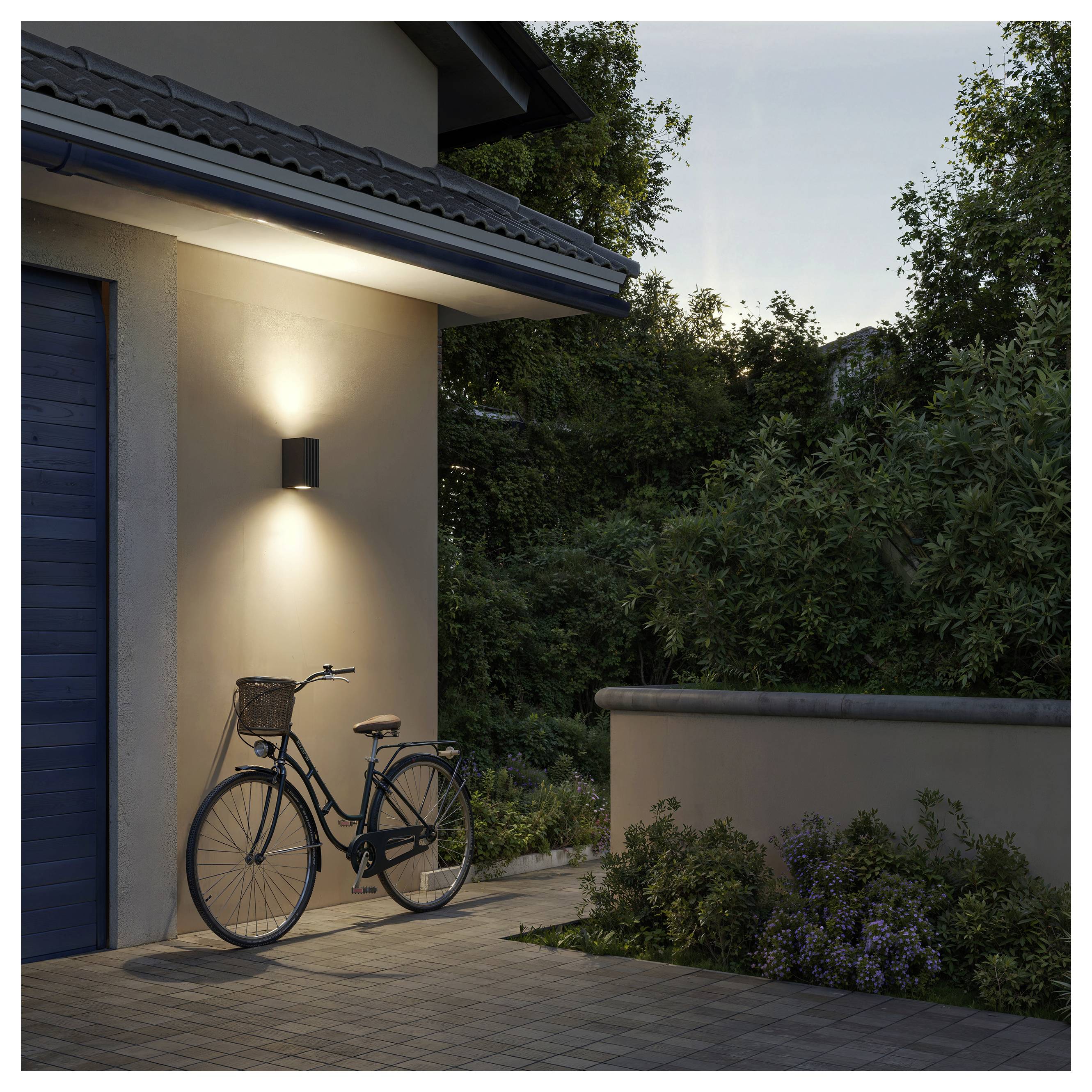 Ein Fahrrad lehnt in der Dämmerung an einer Hauswand unter einer beleuchteten Außenleuchte, neben einer Terrasse und einer Gartenmauer.