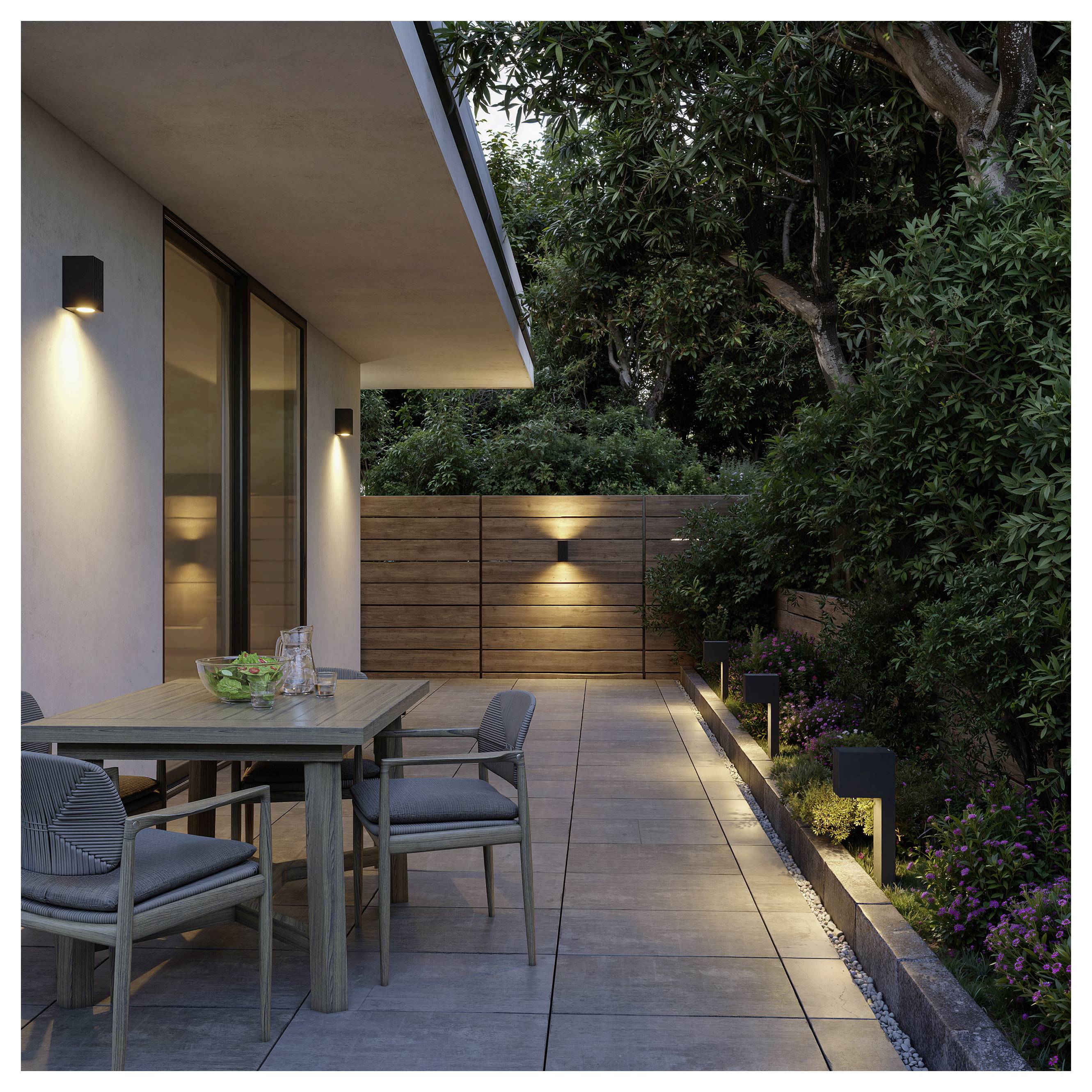 Moderne Terrasse mit Esstisch im Freien, Stühlen und Wandleuchten neben einem Haus, umgeben von Bäumen und angelegtem Garten in der Dämmerung.