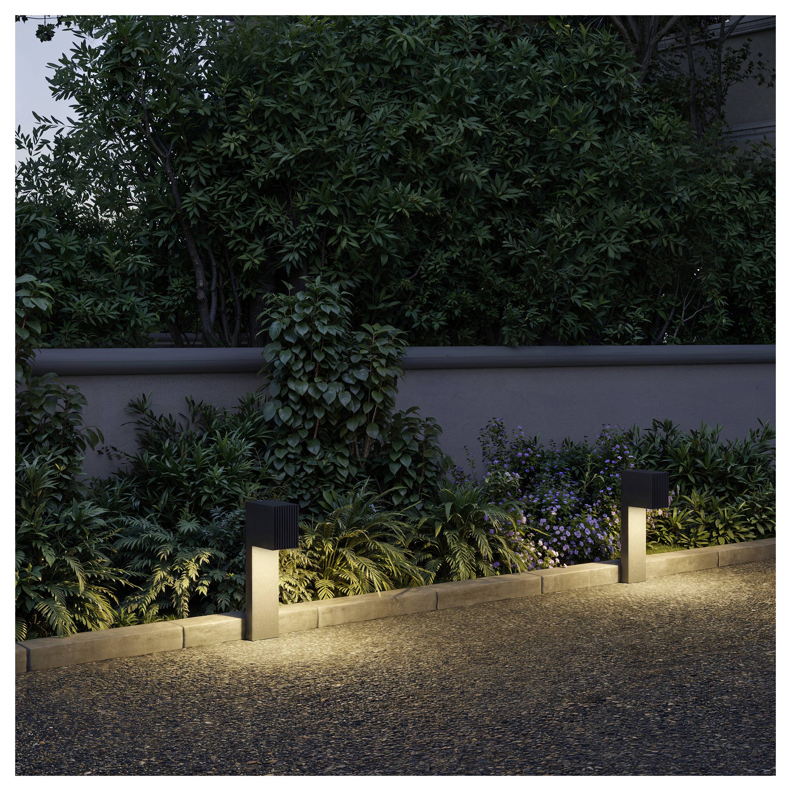 Zwei kleine schwarze Wegleuchten beleuchten einen Gartenborder mit dichten Sträuchern und einer Steinmauer in der Dämmerung.