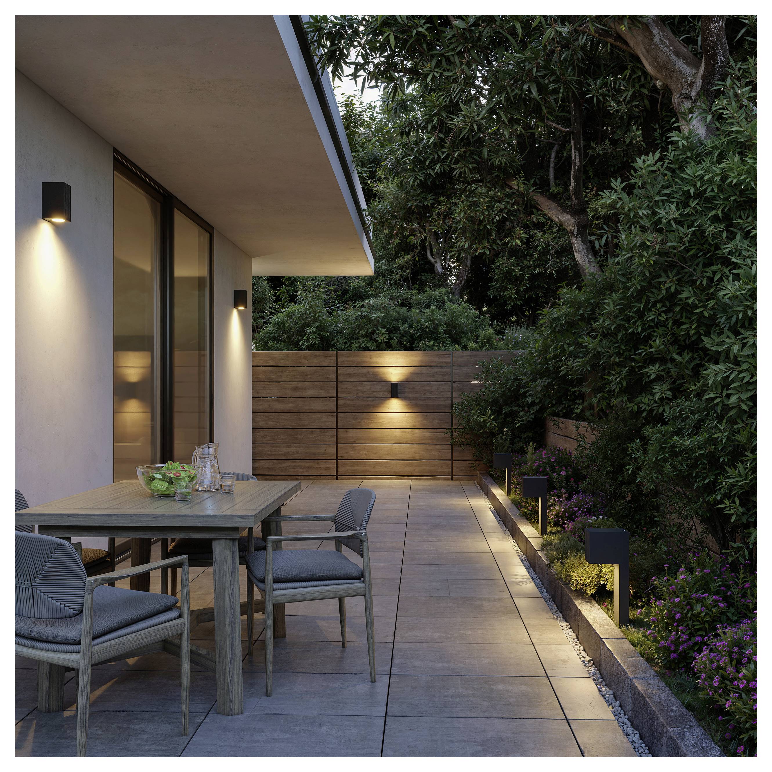 Moderne Terrasse im Freien mit Esstisch und Stühlen neben einem Haus, beleuchtet durch Wand- und Weglichter, umgeben von Holzzaun und üppigen Bäumen.