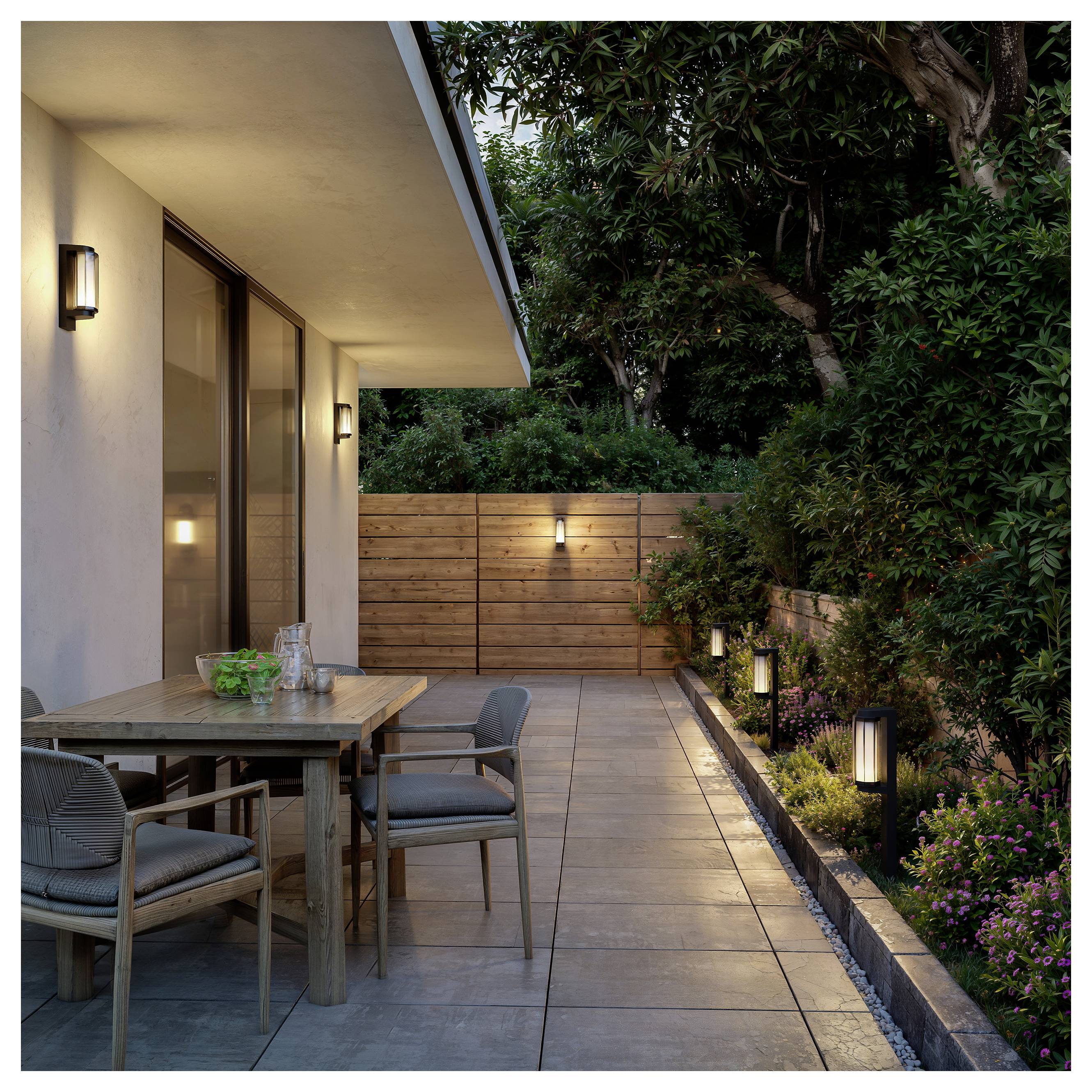 Moderne Terrasse mit Holzesstisch und Stühlen neben einem Haus, umgeben von einem beleuchteten Gartenweg, Holzzaun und hohen Bäumen in der Dämmerung.