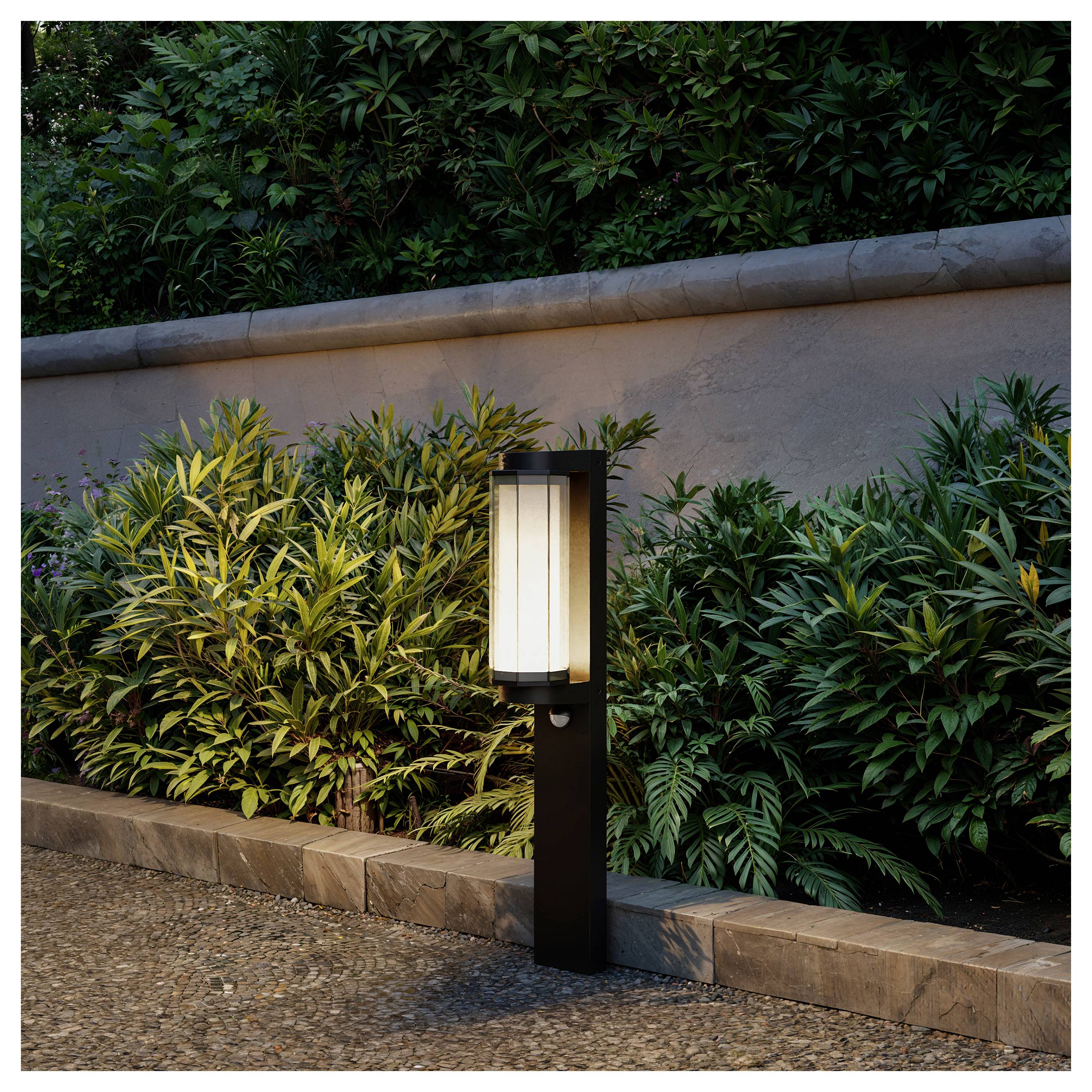 Outdoor schwarzer Poller-Leuchte, die neben einem bepflanzten Blumenbeet und einer Steinmauer in der Dämmerung leuchtet.