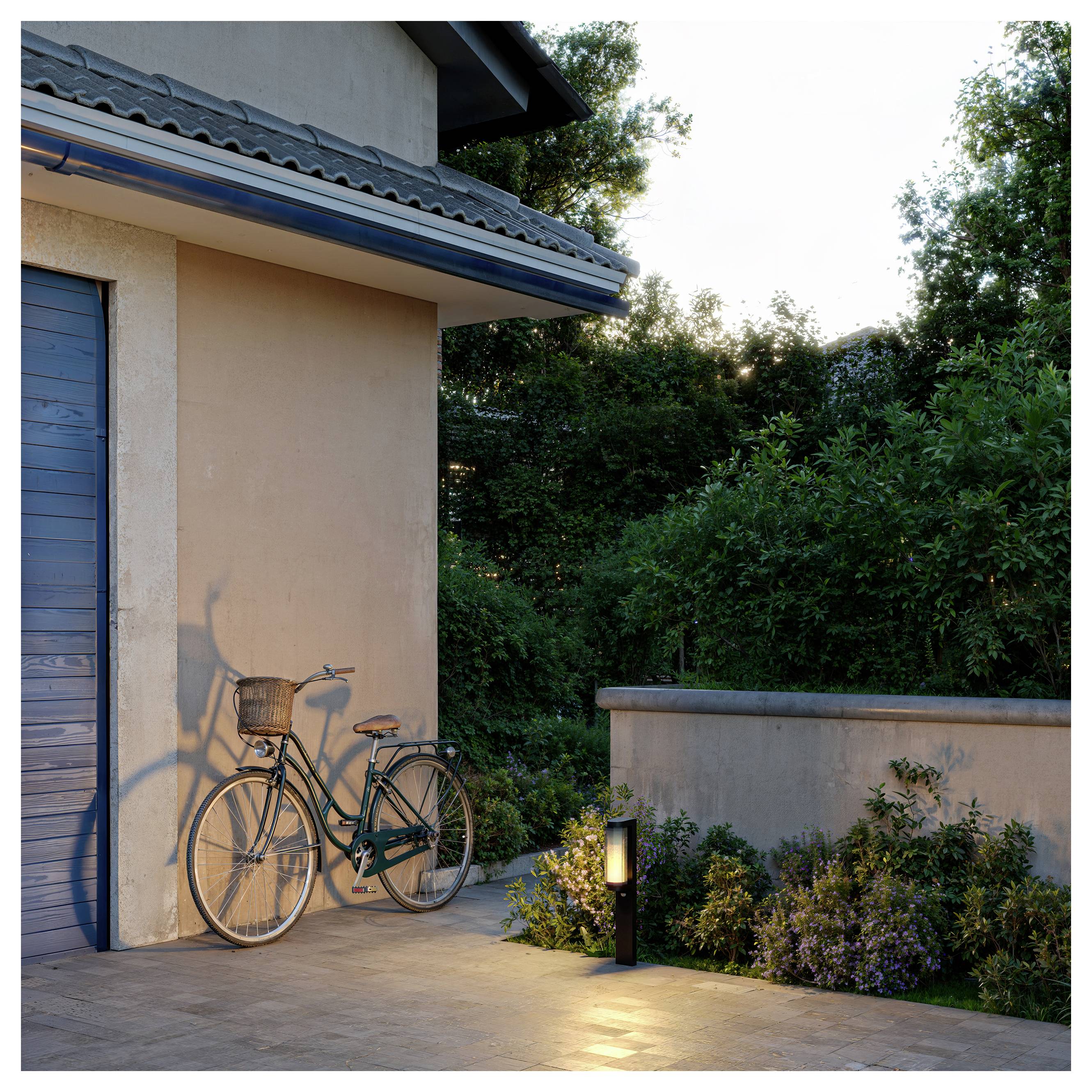 Ein Fahrrad lehnt an einer Hauswand neben einem beleuchteten Gartenweg in der Dämmerung, mit Bäumen und Sträuchern im Hintergrund.