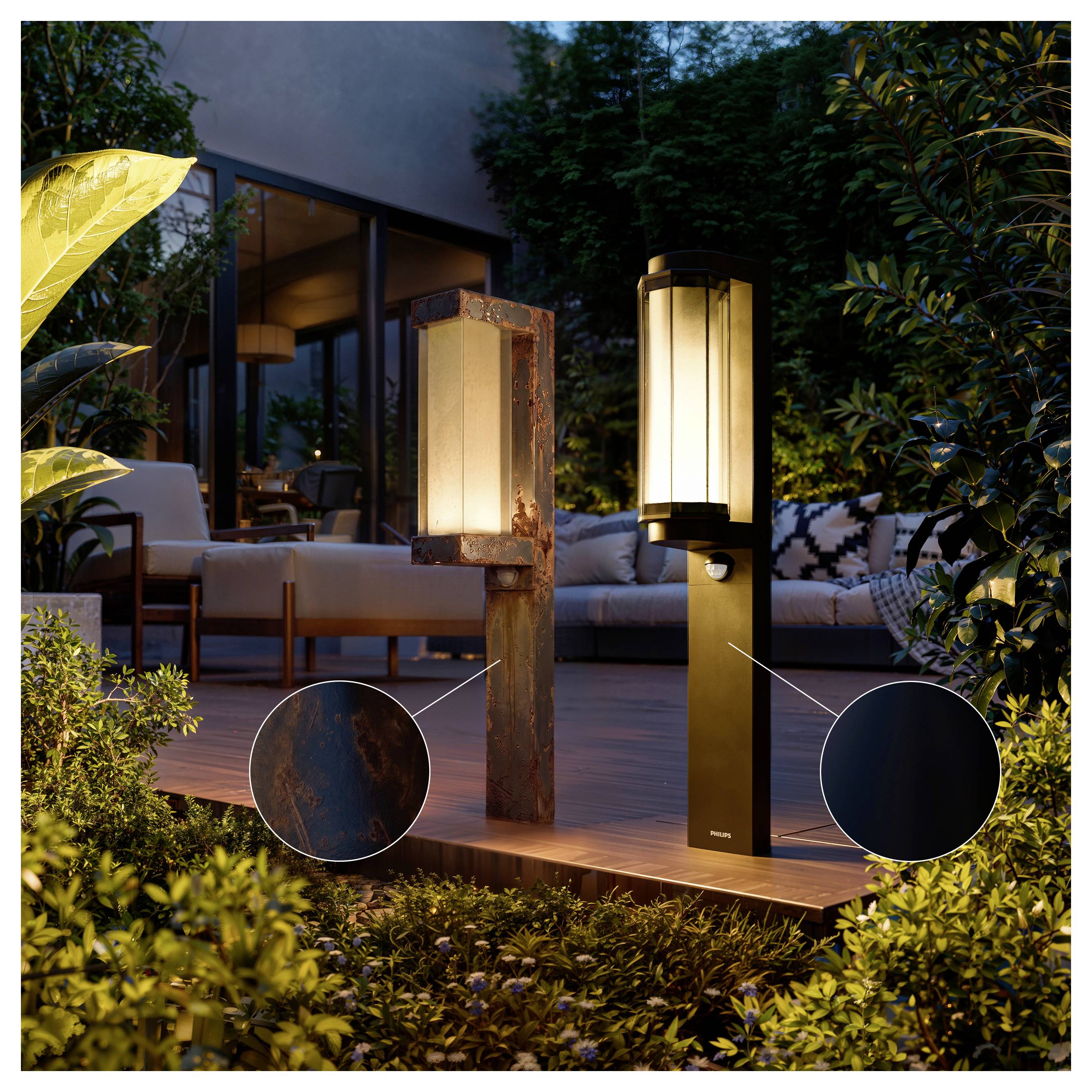 Zwei moderne Außenpfostenleuchten auf einer Terrasse bei Nacht, eine aus verwittertem Metall und eine schwarze, die einen Sitzbereich und einen Garten beleuchten.