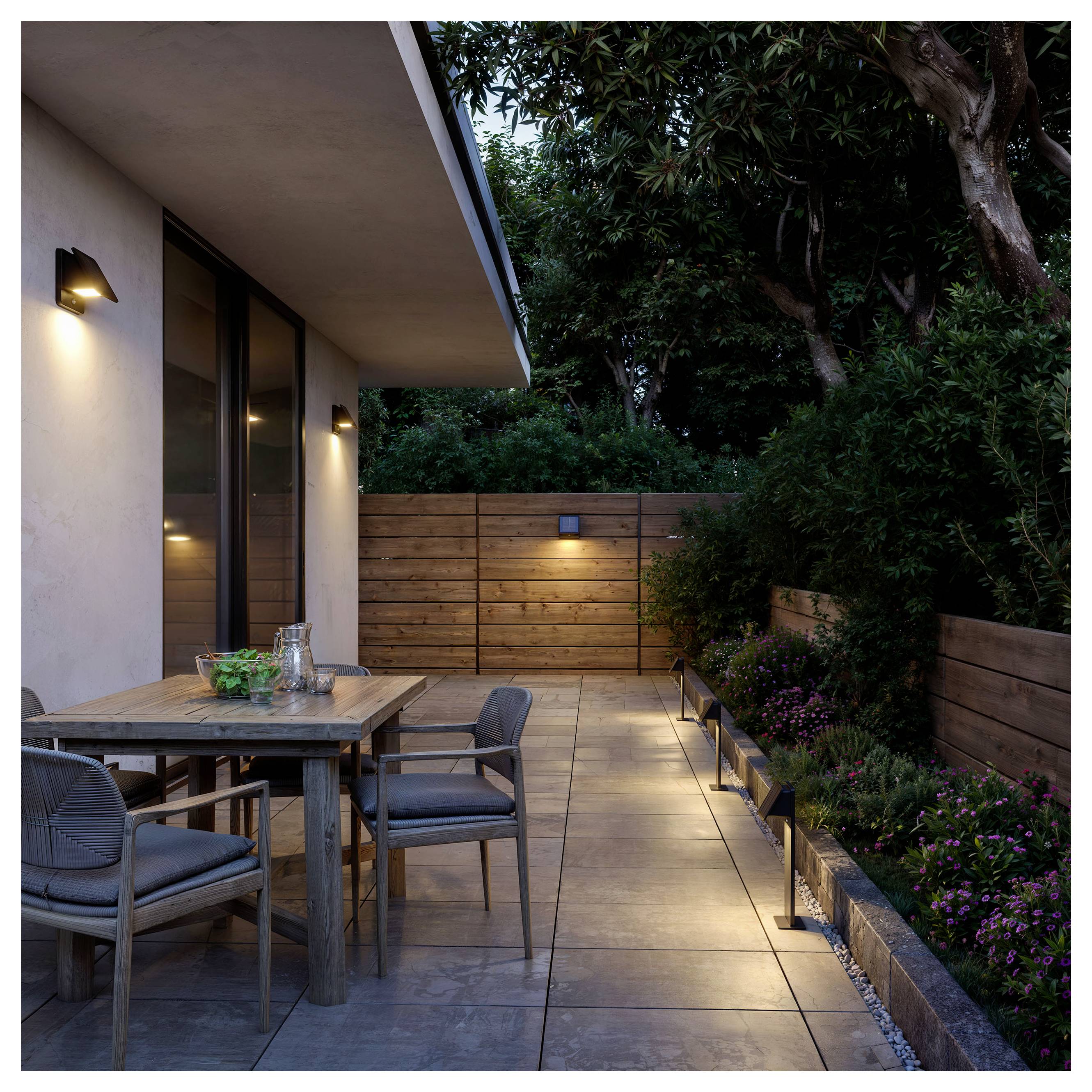 Moderne Terrasse in der Dämmerung mit einem Holzesstisch und Stühlen neben einem Haus, beleuchteter Gehweg und angelegter Garten mit Bäumen und Sträuchern.