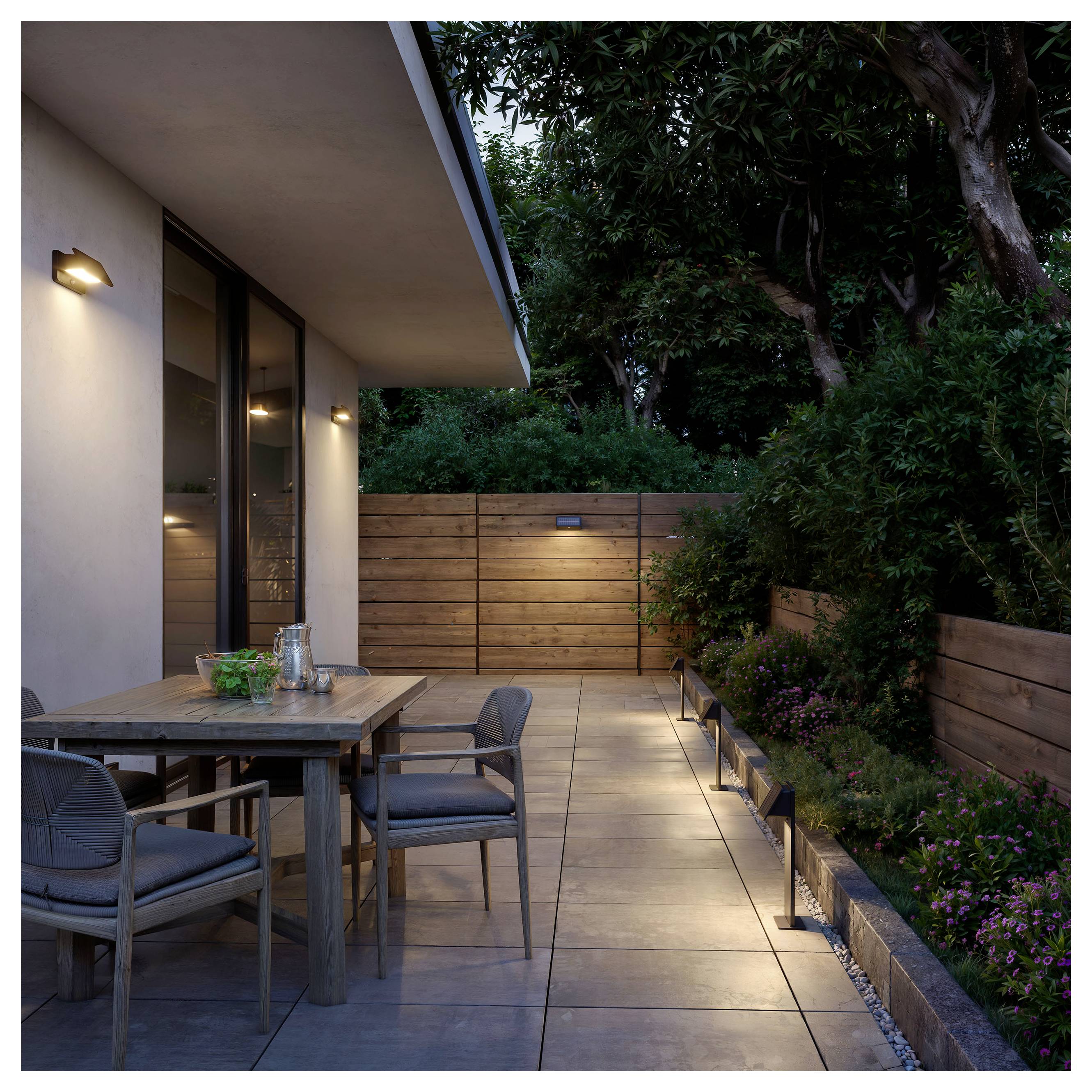 Moderne Terrasse in der Dämmerung mit Esstisch im Freien und Stühlen neben einem Haus, beleuchteter Gehweg, Holzzaun und landschaftlich gestaltete Bäume und Pflanzen.