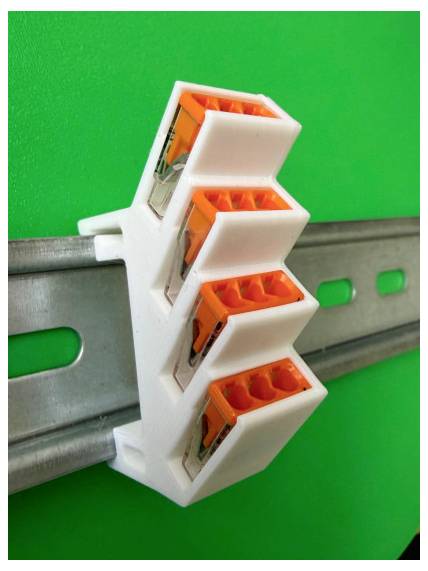 Vier Drahtverbinder mit orangefarbigem Hebel, die in einem weißen Halter montiert sind und an einer Metallschiene vor einem grünen Hintergrund befestigt sind.