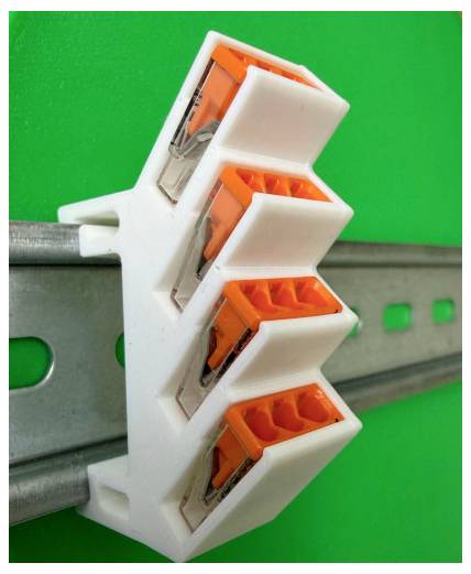 Nahaufnahme einer weißen kunststoffischen Elektroverbinderleiste mit vier orangefarbenen Anschlussöffnungen, die auf einer grauen Metallschiene vor einem grünen Hintergrund montiert ist.