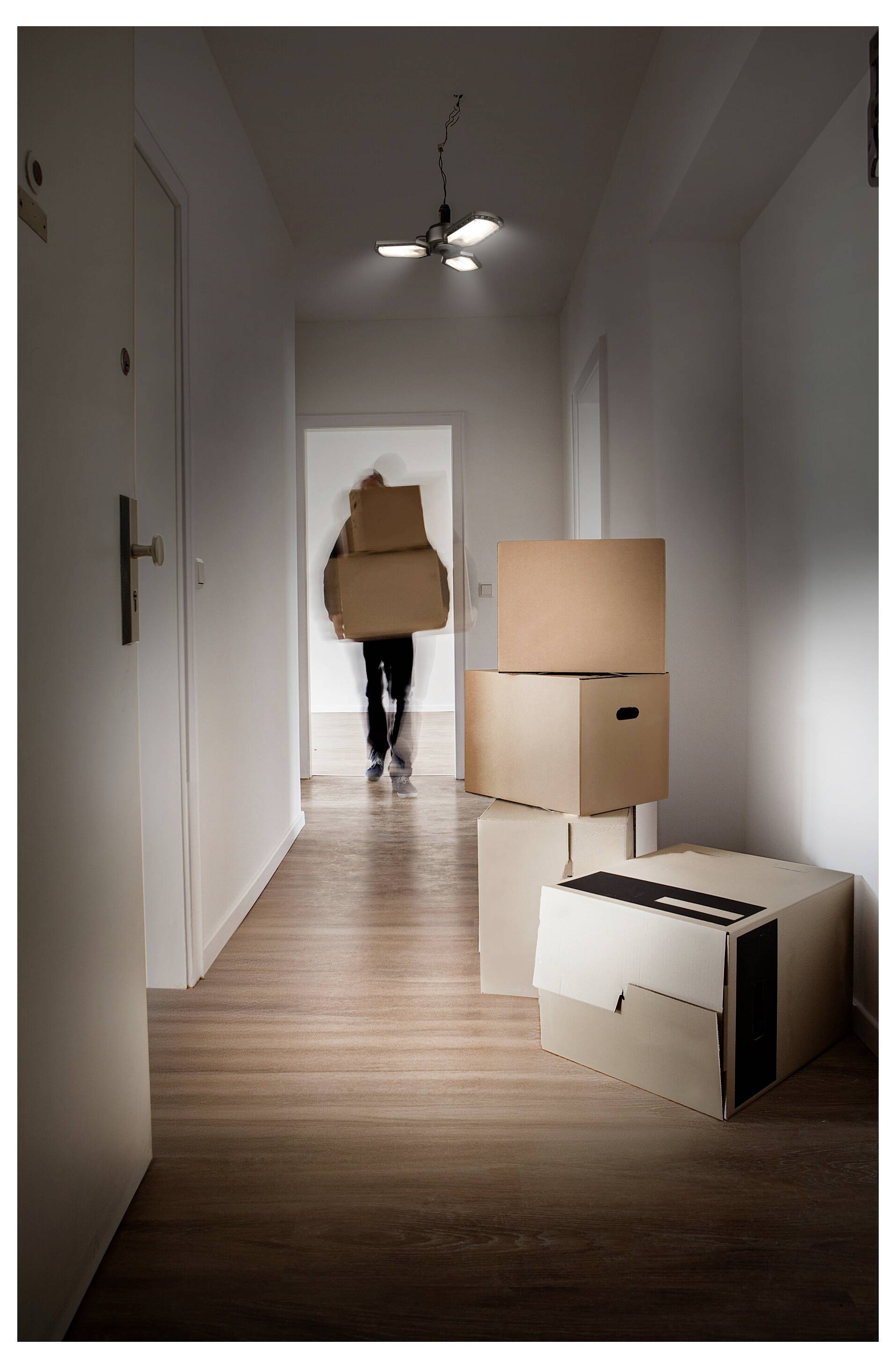 Person, die Umzugskartons einen Flur in einer leeren Wohnung hinunterträgt, mit mehreren gepackten Kartons, die im Vordergrund gestapelt sind.
