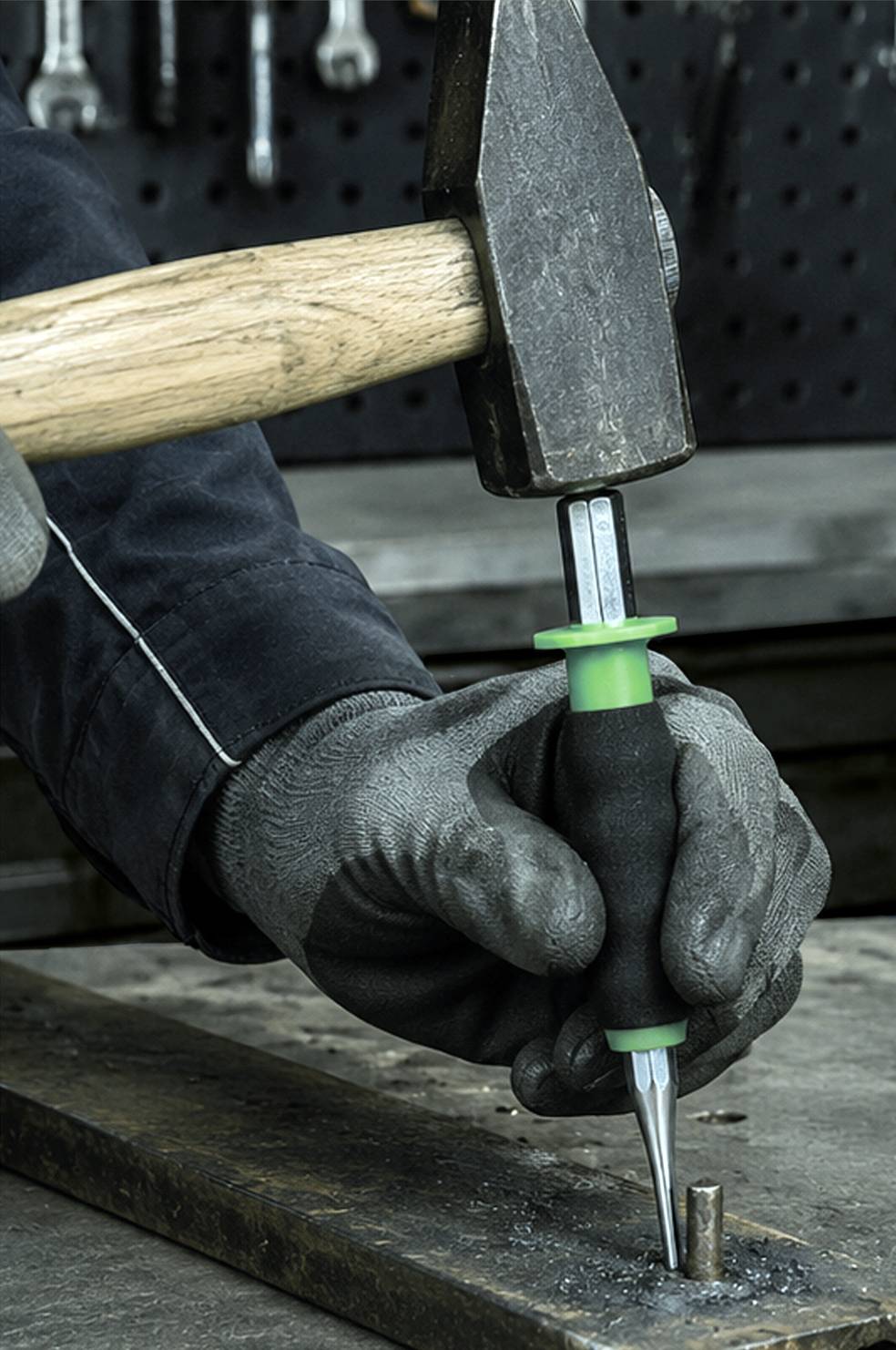 Behandschuhte Hand, die einen Durchschlag unter einem Hammer hält und ihn in eine Metalloberfläche auf einer Werkbank treibt.