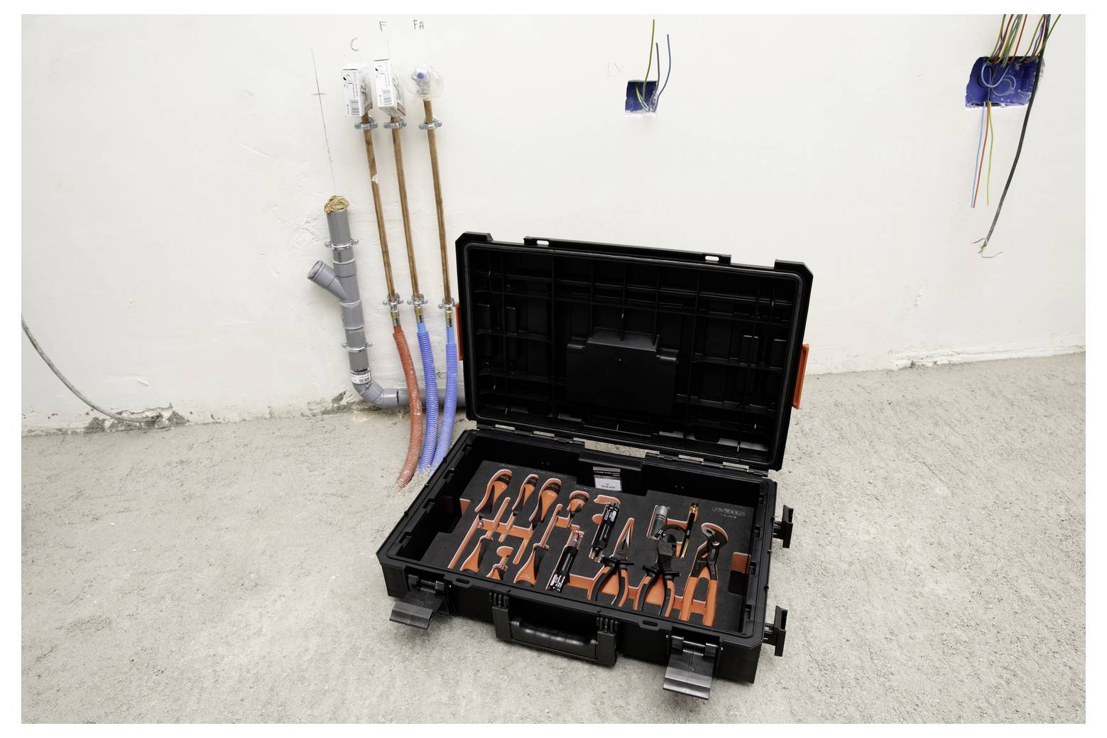 Offener schwarzer Hartschalenkoffer mit orange-griffigen Handwerkzeugen auf einem Betonfußboden, neben wandmontierten Rohren und elektrischen Leitungen in einem unvollendeten Raum.